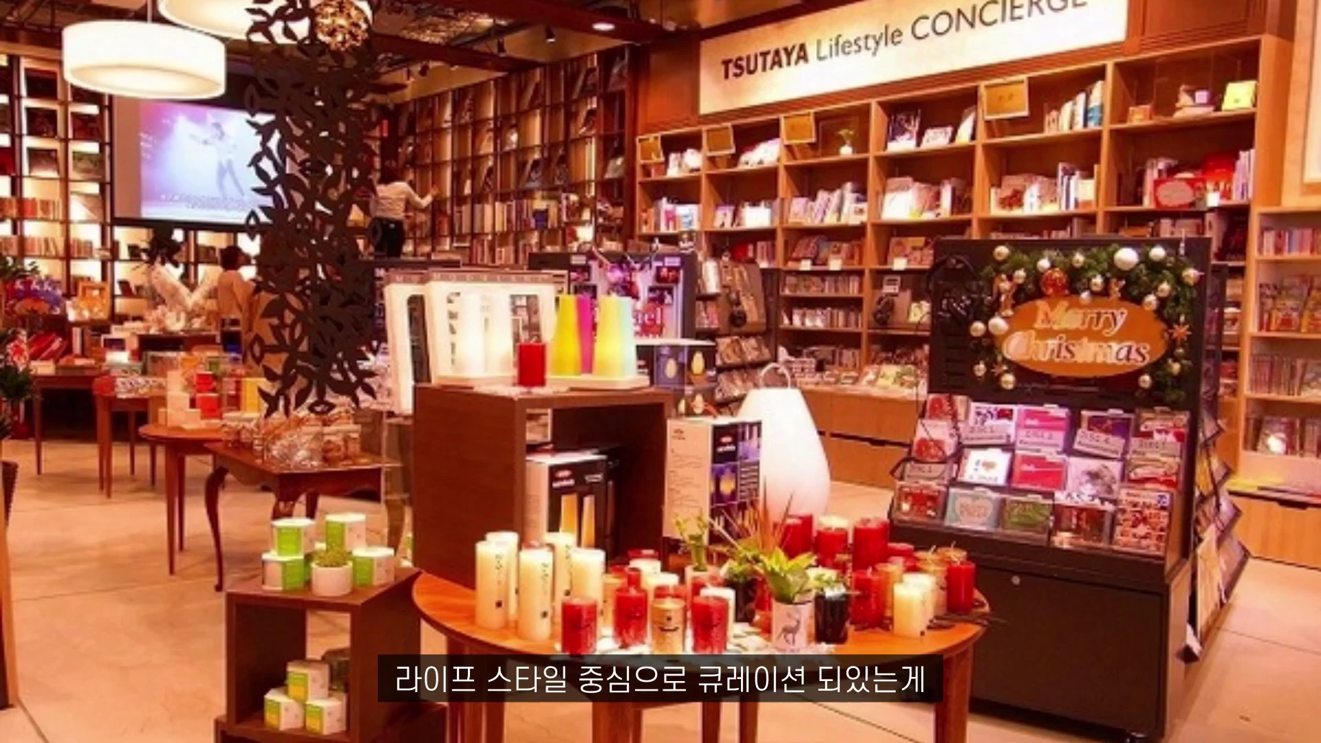 나무 선반에 책과 소품이 진열된 라이프스타일 서점 내부 모습으로 중앙 원형 테이블 위에 양초와 식물 등이 놓여 있고 벽면에는 'TSUTAYA Lifestyle CONCIERGE'라는 문구가 보입니다.