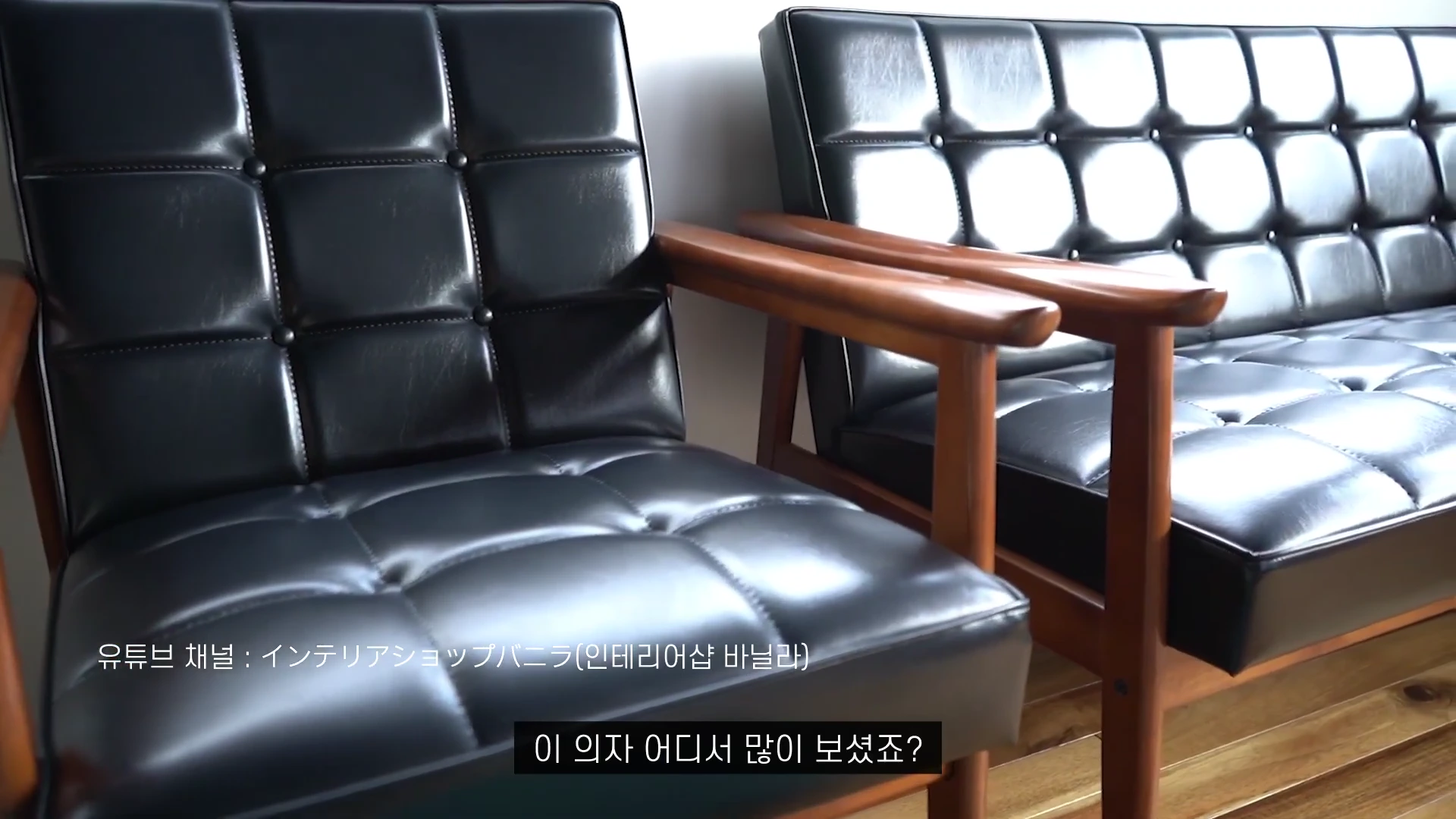 나무 팔걸이가 있는 검은색 가죽 소파 두 개가 나란히 놓여 있고 화면 하단에는 '이 의자 어디서 많이 보셨죠?'라는 자막이 적혀 있다.