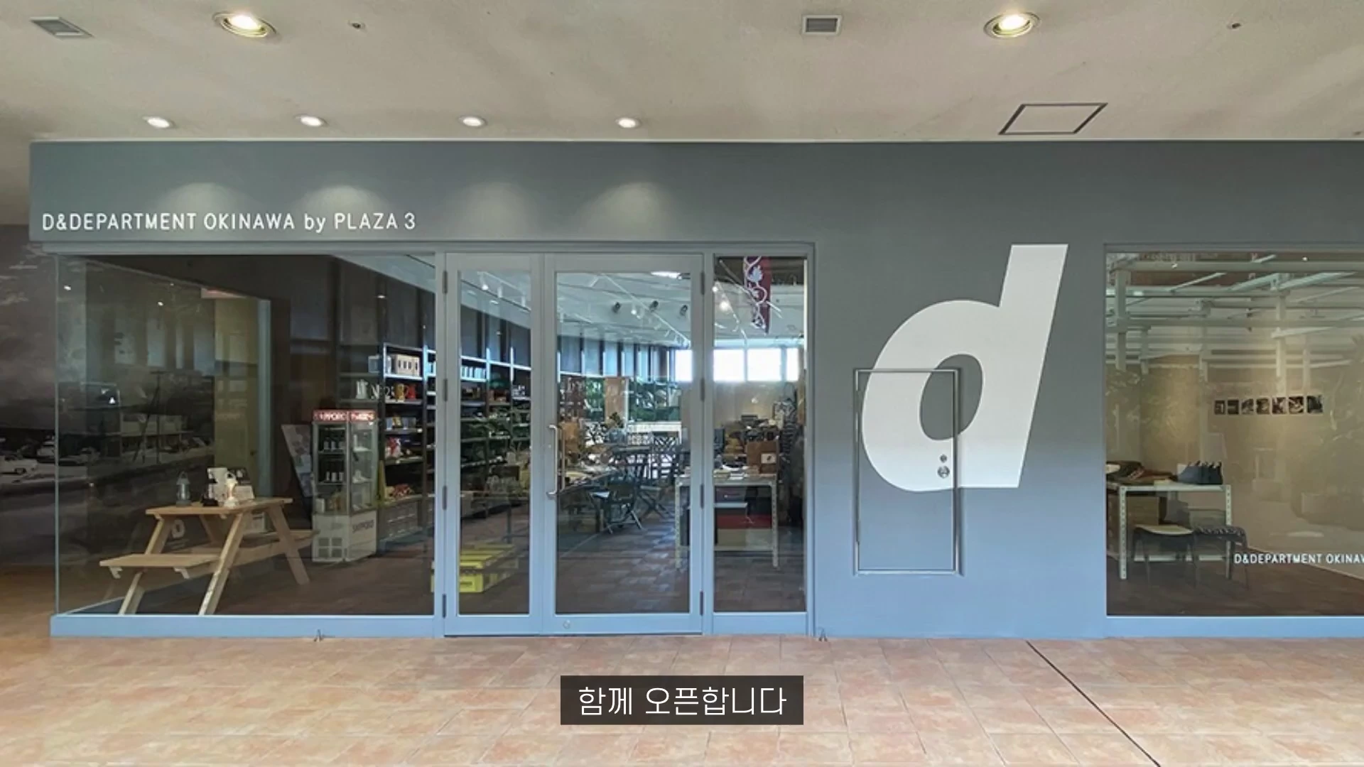 D&D 오키나와 바이 플라자 3 매장의 정면 모습으로, 회색 외벽에 커다란 흰색 'd' 로고가 그려져 있고 유리창을 통해 내부의 가구와 진열대가 보입니다.