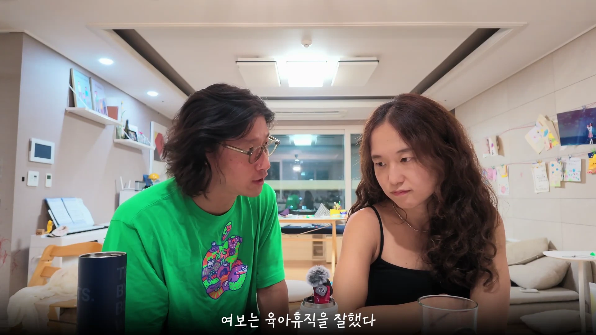 긴 머리의 남성과 여성이 실내에서 서로를 바라보며 대화를 나누고 있는 모습.