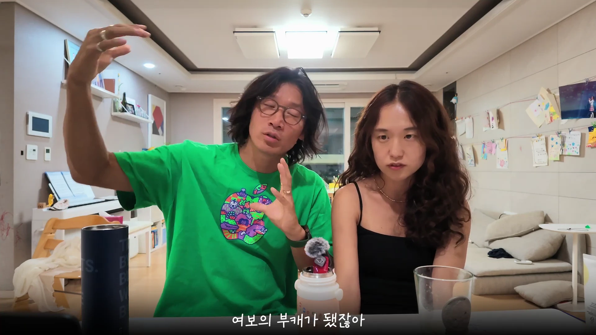 실내 거실을 배경으로 초록색 티셔츠를 입은 남성과 검은색 민소매 옷을 입은 여성이 나란히 앉아 대화하고 있는 모습이다.