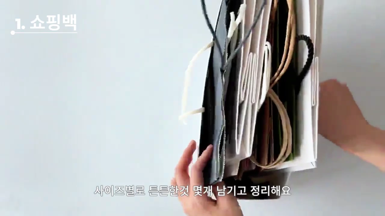 흰색 배경 위에서 양손으로 다양한 크기의 쇼핑백들을 정리하고 있는 모습이며, 화면 상단에 '1. 쇼핑백'이라는 문구와 하단에 자막이 표시되어 있습니다.