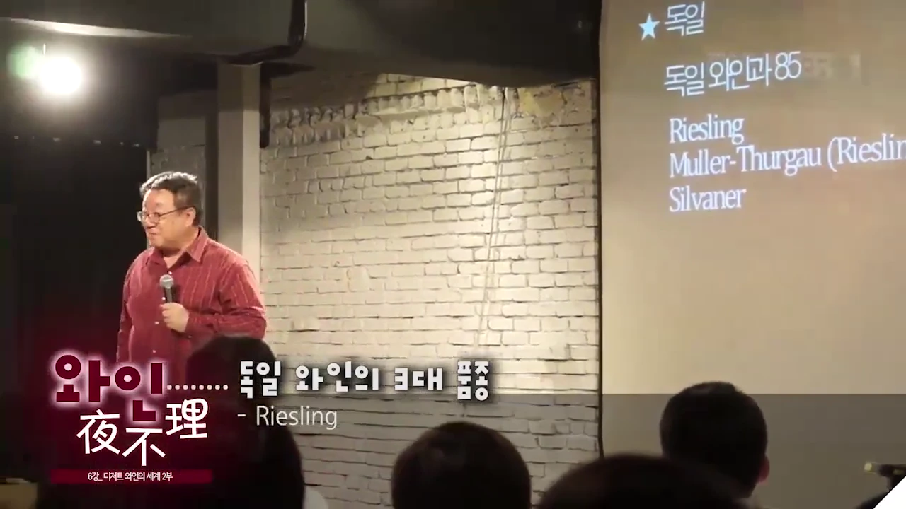 강연장에서 한 남성이 마이크를 잡고 서 있고, 옆 대형 스크린에는 독일 와인 품종 목록이 적힌 슬라이드가 비치고 있다.