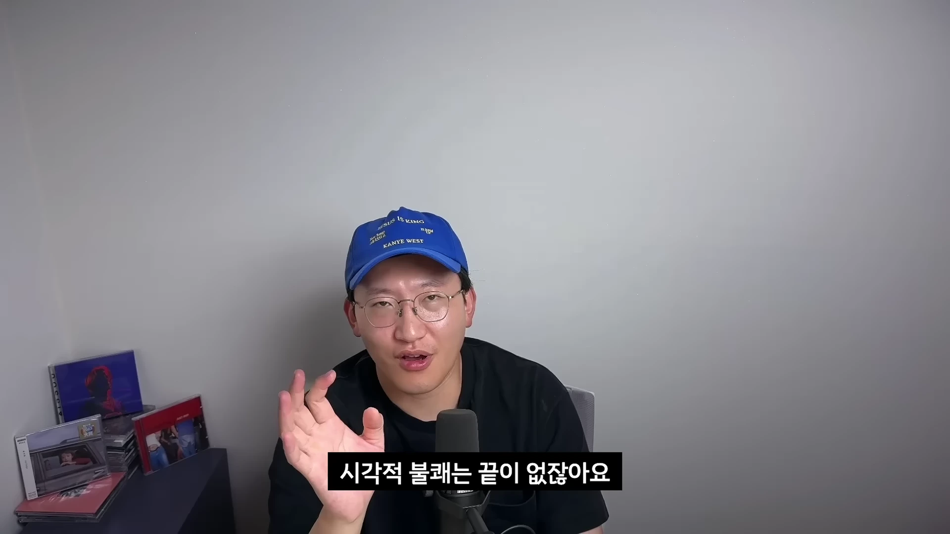 파란색 볼캡 모자를 쓰고 안경을 쓴 남성이 마이크 앞에서 손동작을 하며 이야기하고 있으며 화면 하단에는 '시각적 불쾌는 끝이 없잖아요'라는 자막이 표시되어 있습니다.