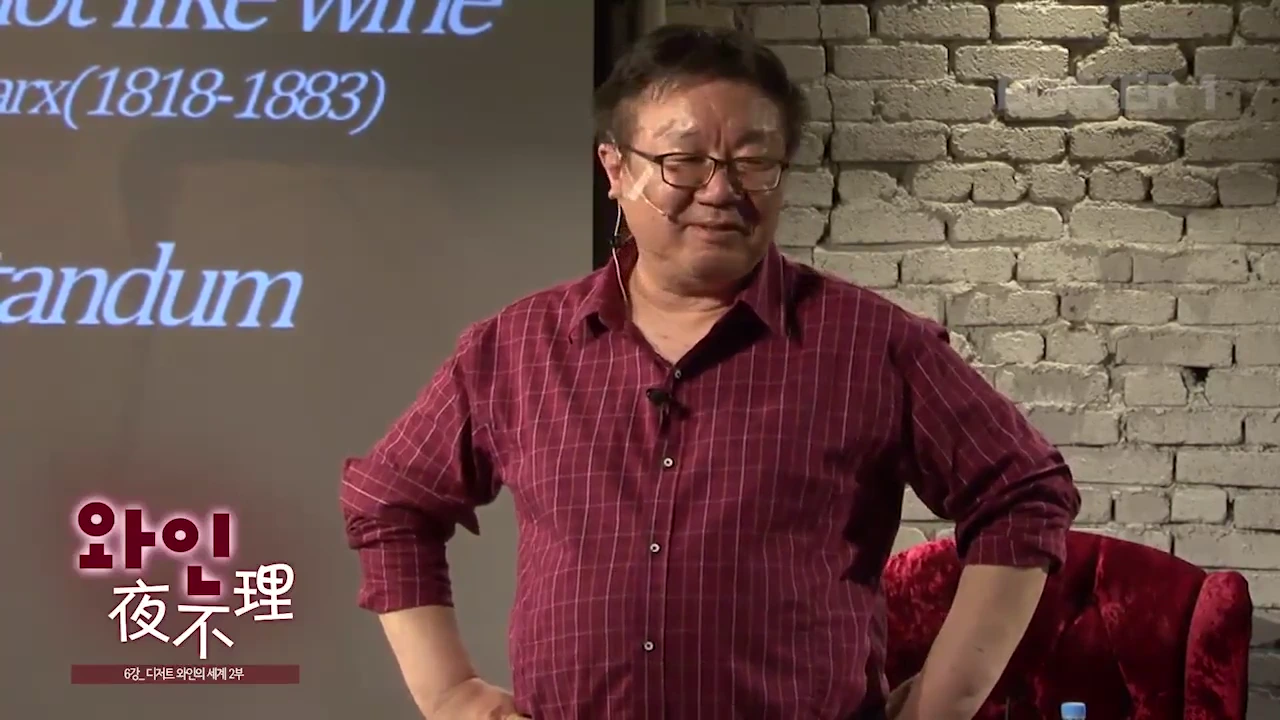 안경을 쓴 중년 남성이 벽돌 벽을 배경으로 무대 위에 서서 청중을 바라보며 이야기하고 있고, 뒤편 화면에는 카를 마르크스 관련 문구가 희미하게 비치고 있습니다.