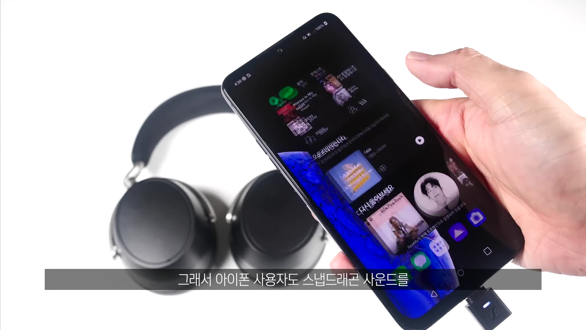 검은색 헤드폰 옆에서 손으로 스마트폰을 들고 있는 모습이며, 스마트폰 하단에는 작은 동글이 연결되어 있습니다.