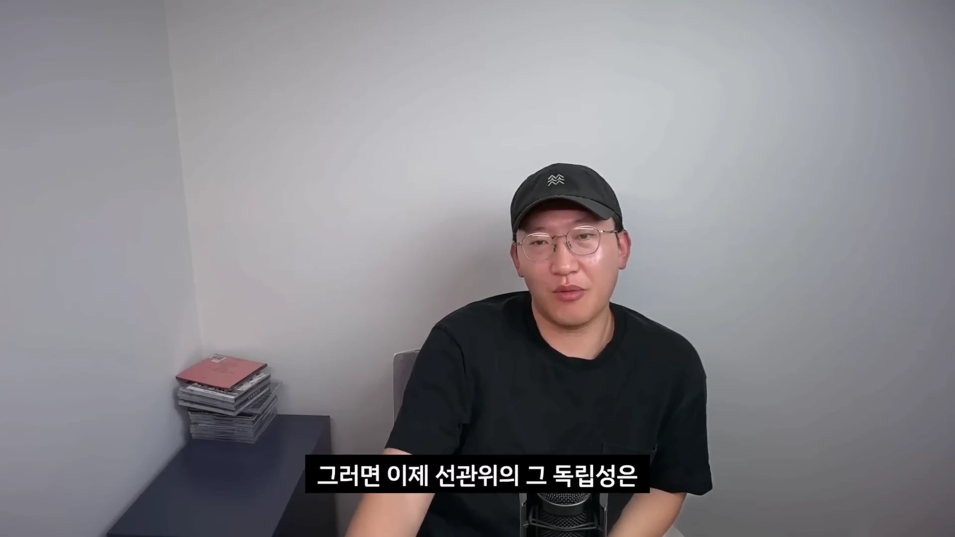 안경을 쓰고 검은색 야구 모자와 티셔츠를 입은 남성이 흰 벽을 배경으로 앉아 이야기를 나누고 있는 모습