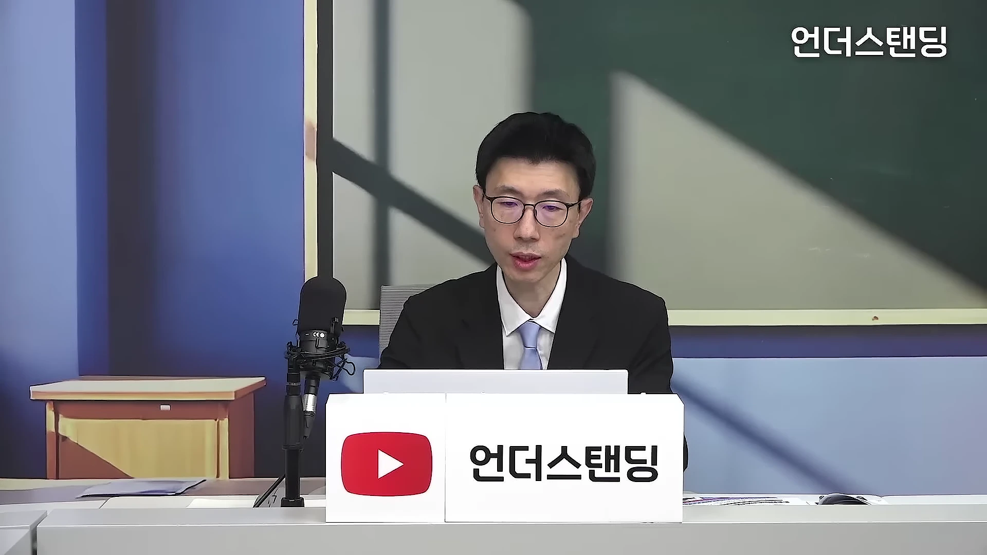 안경을 쓴 남성 진행자가 마이크가 설치된 스튜디오 책상 앞에 앉아 이야기하고 있는 모습