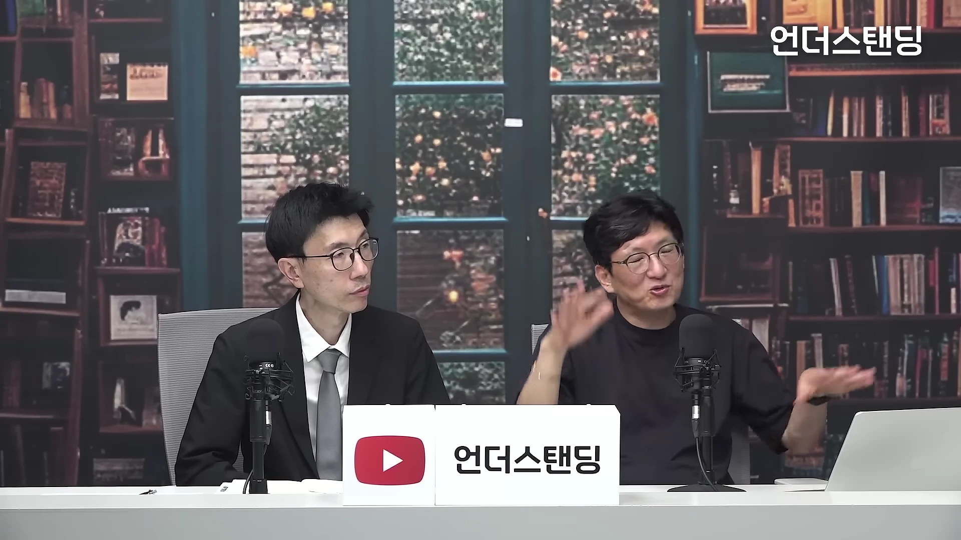 서재 배경의 스튜디오에서 안경을 쓴 두 명의 남성이 각자의 마이크 앞에 앉아 대화를 나누고 있다.