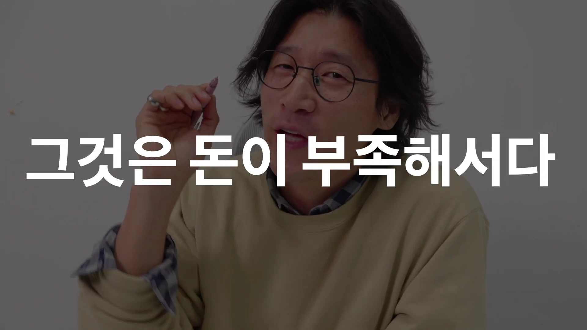 안경을 쓴 남성이 손에 펜을 들고 카메라를 응시하고 있으며 화면 중앙에는 '그것은 돈이 부족해서다'라는 문구가 흰색 글씨로 적혀 있다.