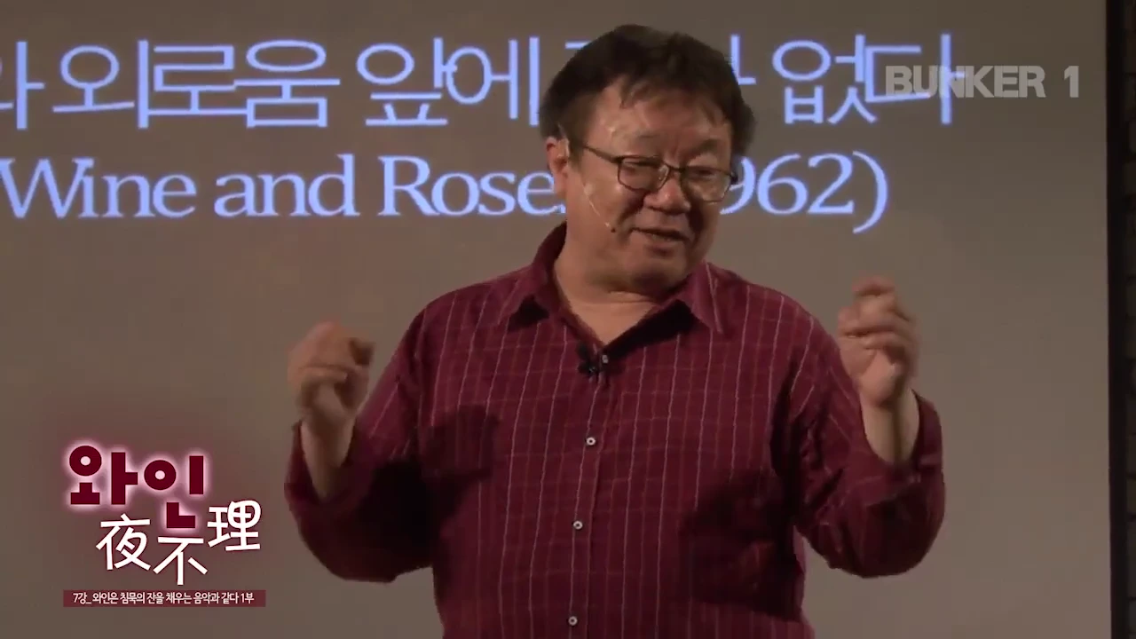 붉은 체크무늬 셔츠를 입고 안경을 쓴 중년 남성이 강의실 강단에서 손동작을 하며 이야기를 하고 있고, 뒤편 화면에는 '외로움 앞에'와 'Wine and Roses'라는 문구가 일부 표시되어 있습니다.