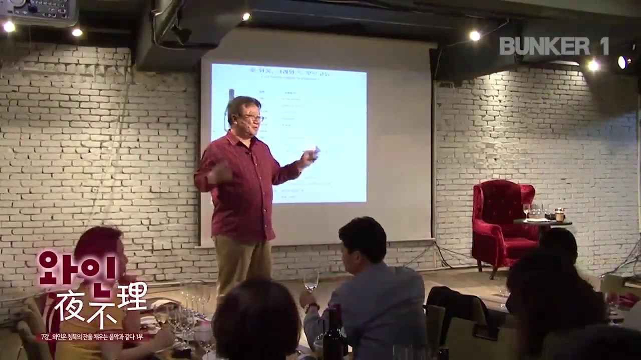 강단에서 와인에 대해 강의하는 발표자와 그 앞에 앉아 경청하는 청중들이 있는 실내 강연장 모습.