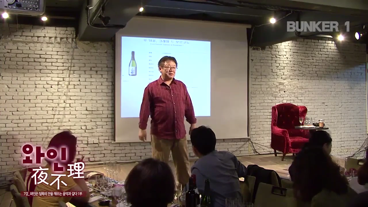 실내 강연장에서 강연자가 흰색 벽면 스크린 앞에 서서 청중을 향해 이야기하고 있으며, 스크린에는 루 디몽 크레망 드 부르고뉴 와인 병 사진과 관련 정보가 투사되어 있습니다.