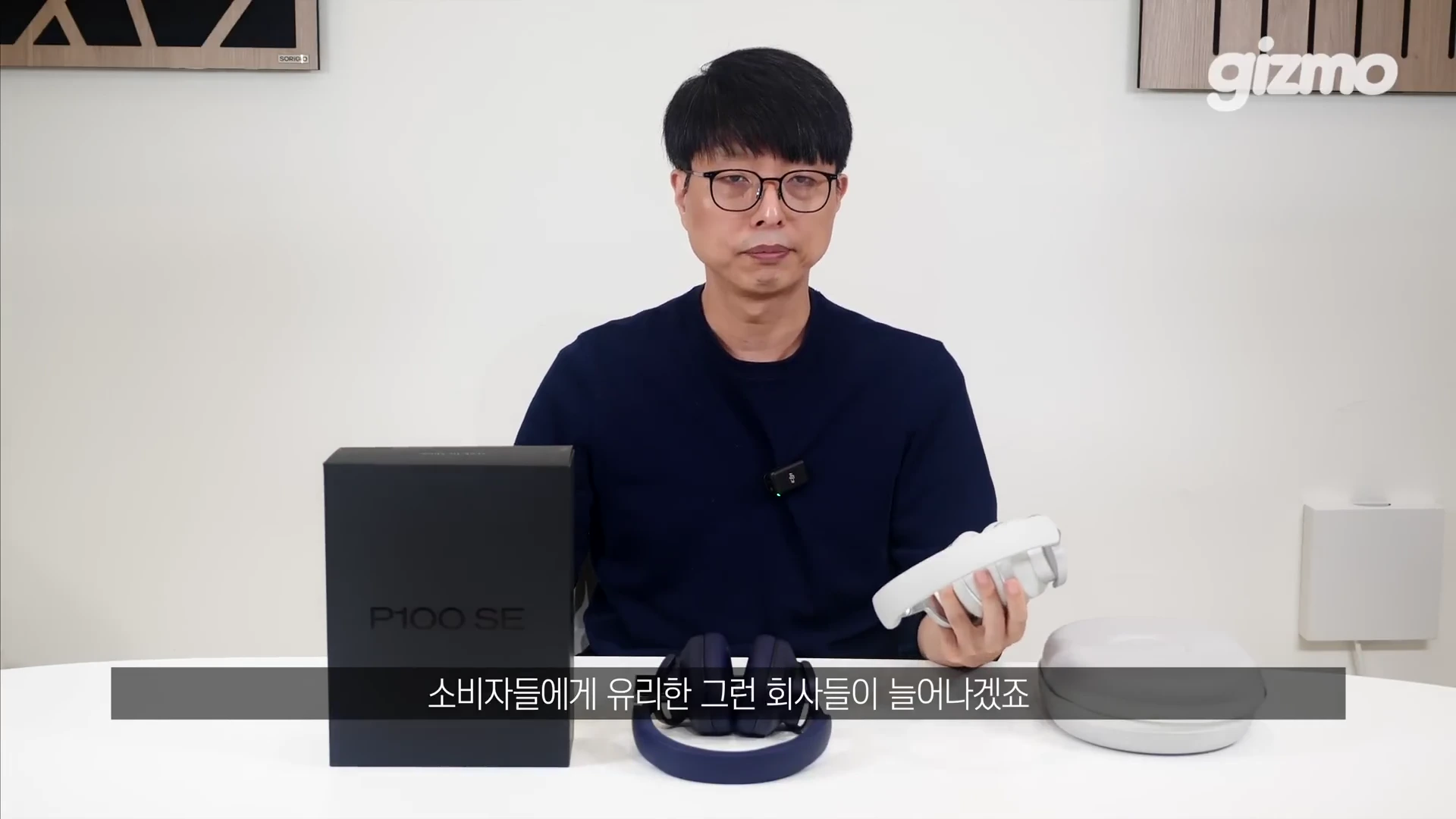 안경을 쓴 남성이 책상 앞에 앉아 한 손에는 흰색 무선 헤드폰을 들고, 앞에는 검은색 제품 상자와 파란색 헤드폰, 그리고 케이스를 놓고 설명하는 모습