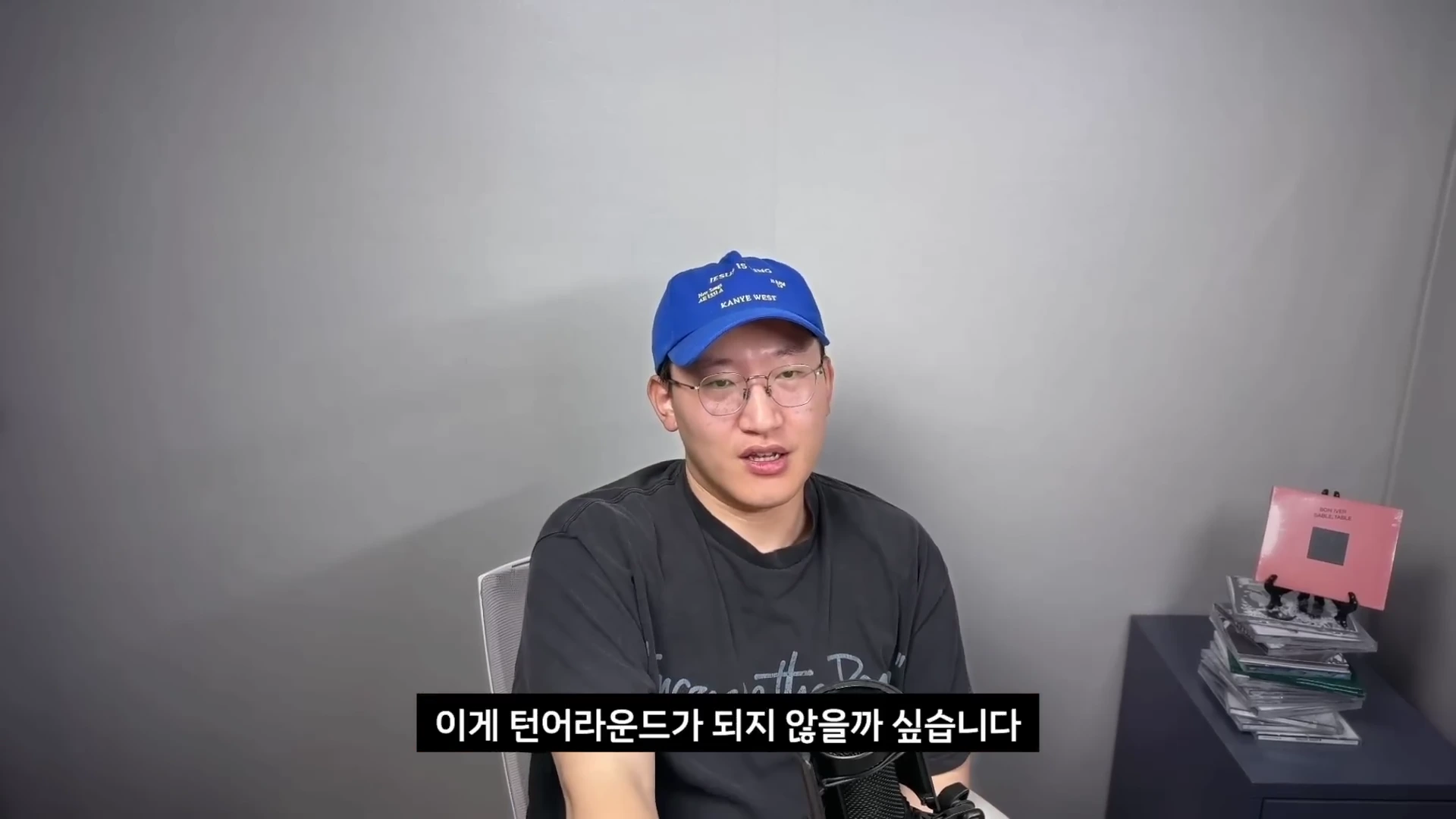 파란색 야구 모자와 안경을 착용한 남성이 회색 배경의 실내에서 마이크 앞에 앉아 이야기하는 모습