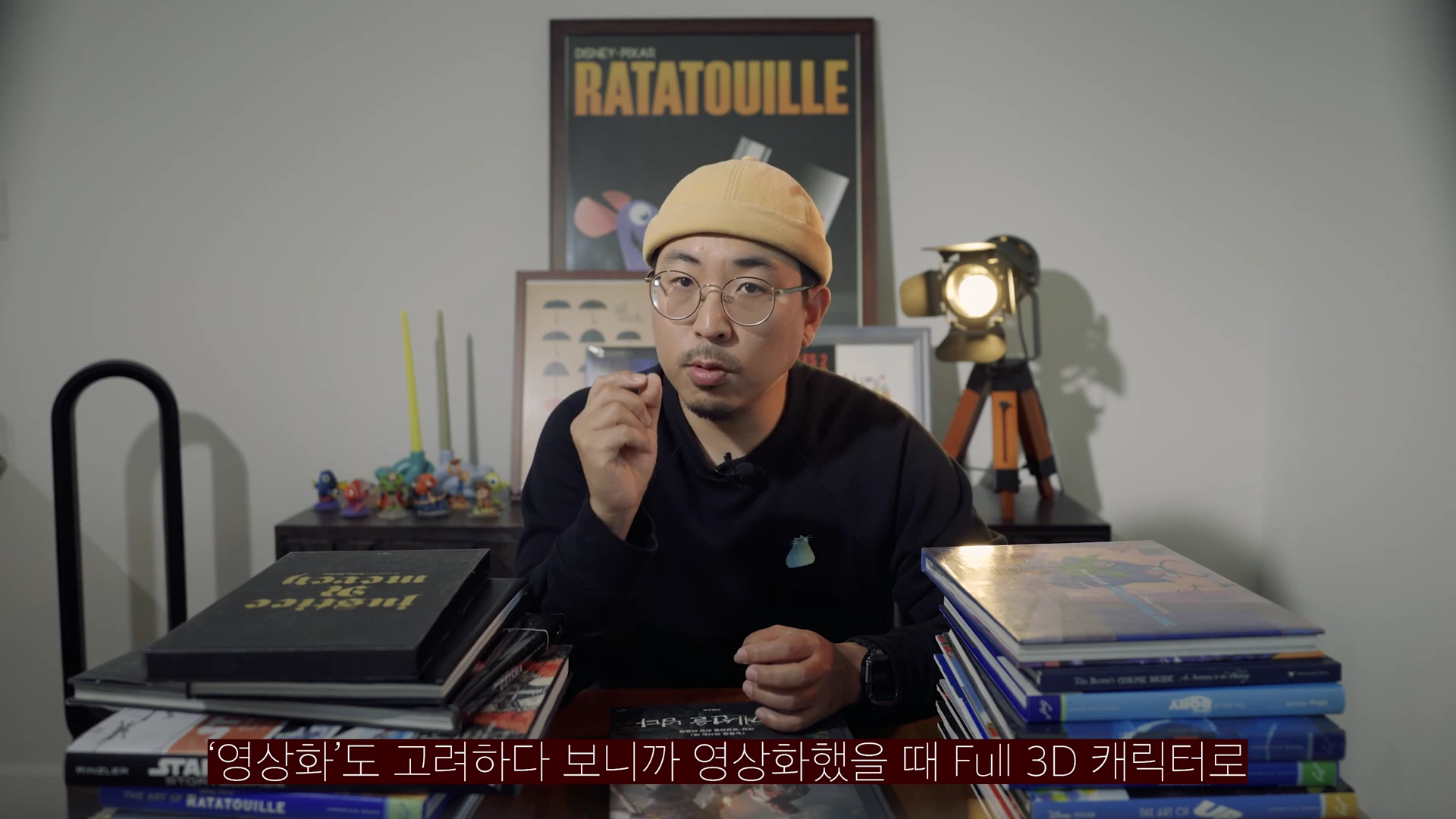 노란색 비니를 쓴 안경을 쓴 남성이 책상이 놓인 실내에서 손짓하며 이야기를 나누고 있다.
