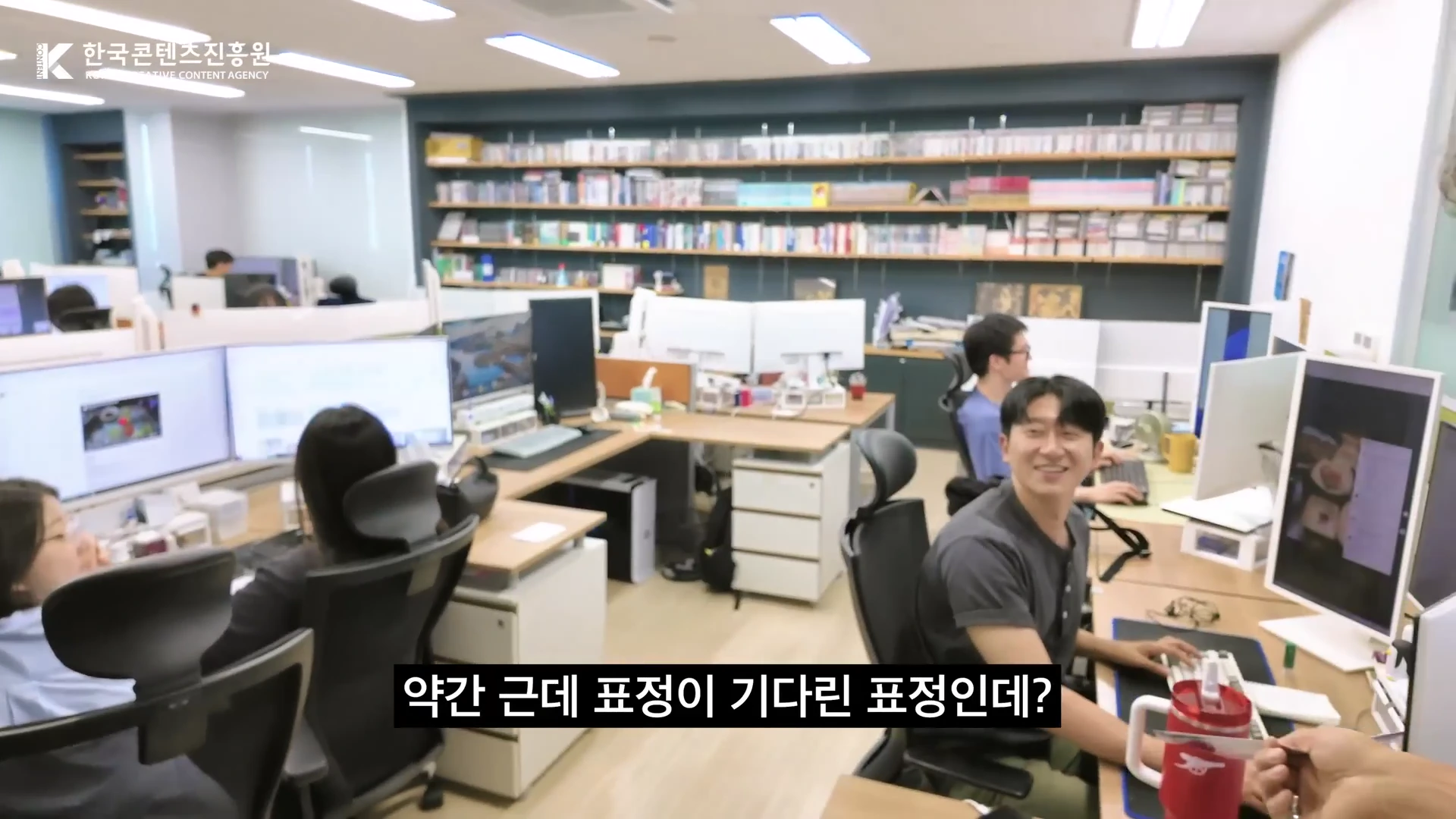 사무실 내에서 여러 명의 직원이 각자의 책상에 앉아 모니터를 보고 일하는 모습이며, 앞쪽에 앉은 남성 직원이 카메라를 향해 웃고 있다.