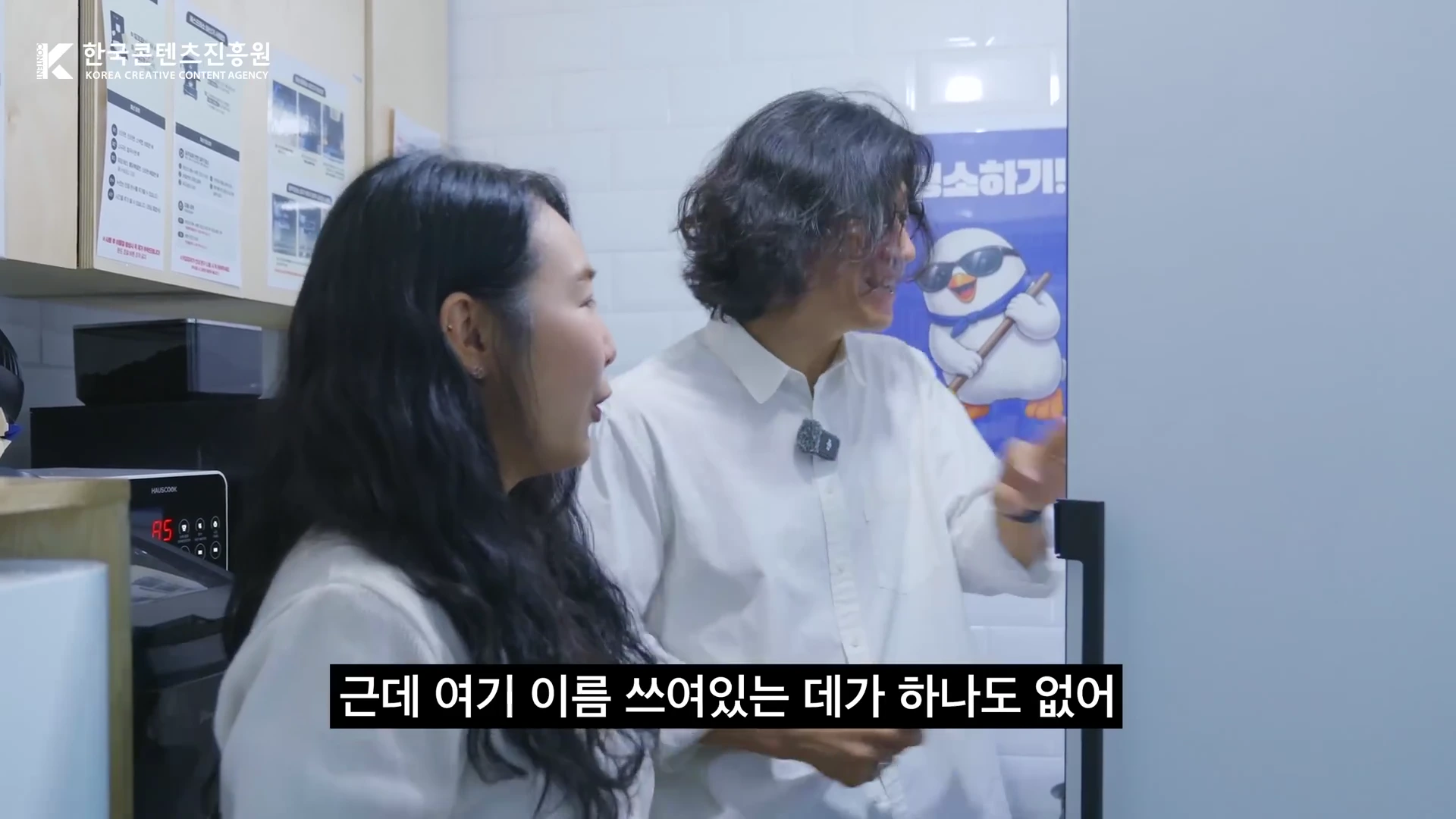 흰 셔츠를 입은 두 사람이 사무실 탕비실 앞에서 냉장고를 보며 대화를 나누고 있다.