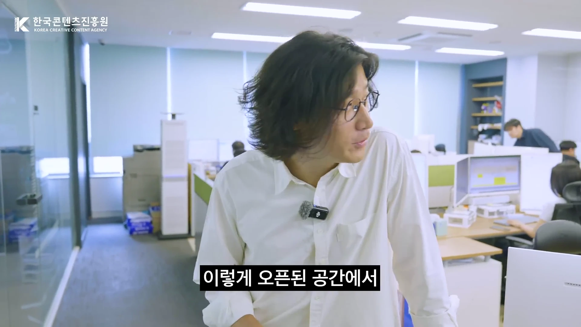 안경을 쓴 남성이 사무실 공간에 서서 이야기를 나누고 있으며, 배경으로 파티션이 나누어진 사무 공간과 업무 중인 사람들이 보인다.