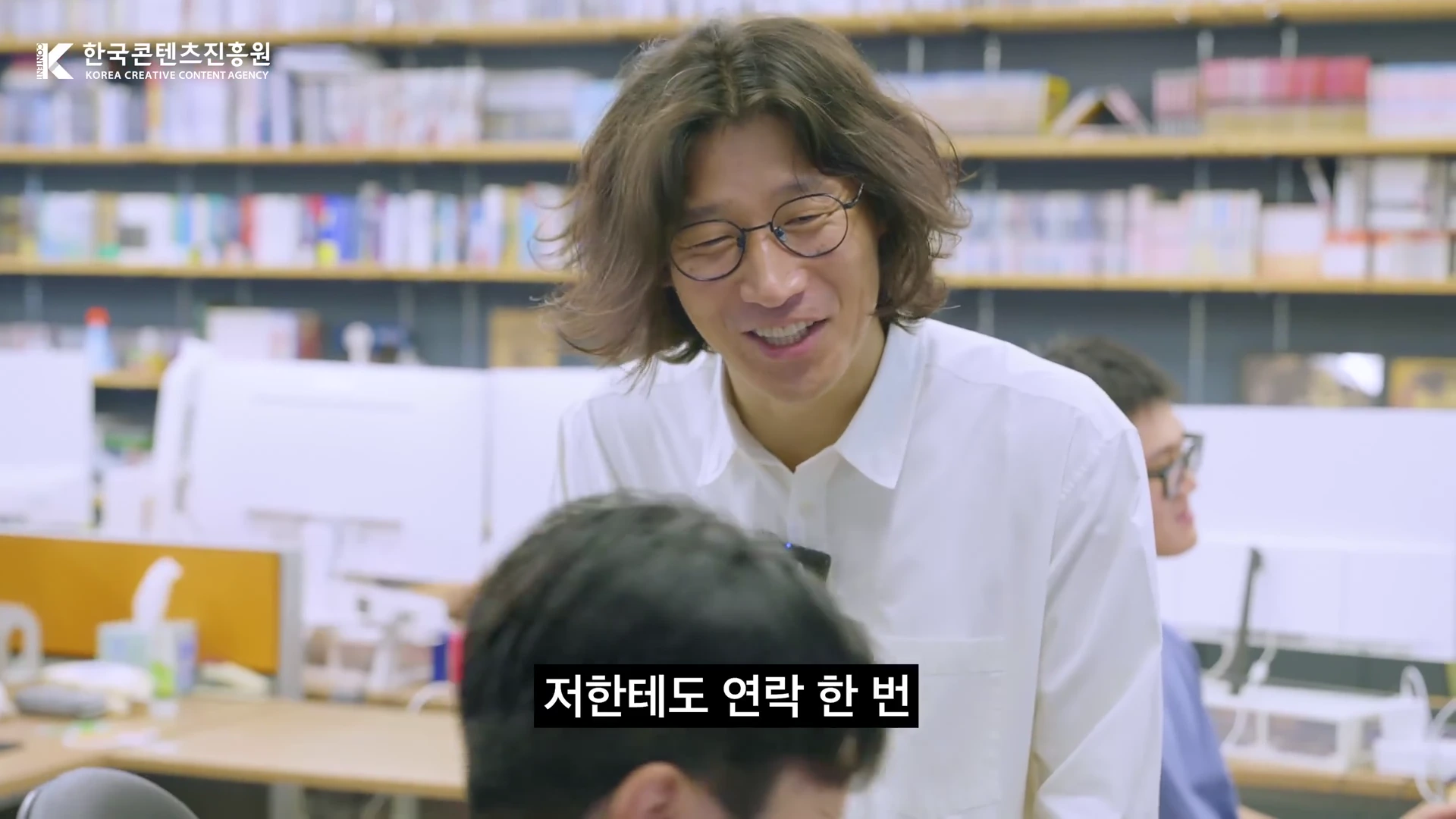 안경을 쓴 긴 머리 남성이 사무실 책상에 앉아 있는 직원에게 다가가 웃으며 대화를 나누고 있는 모습이다.