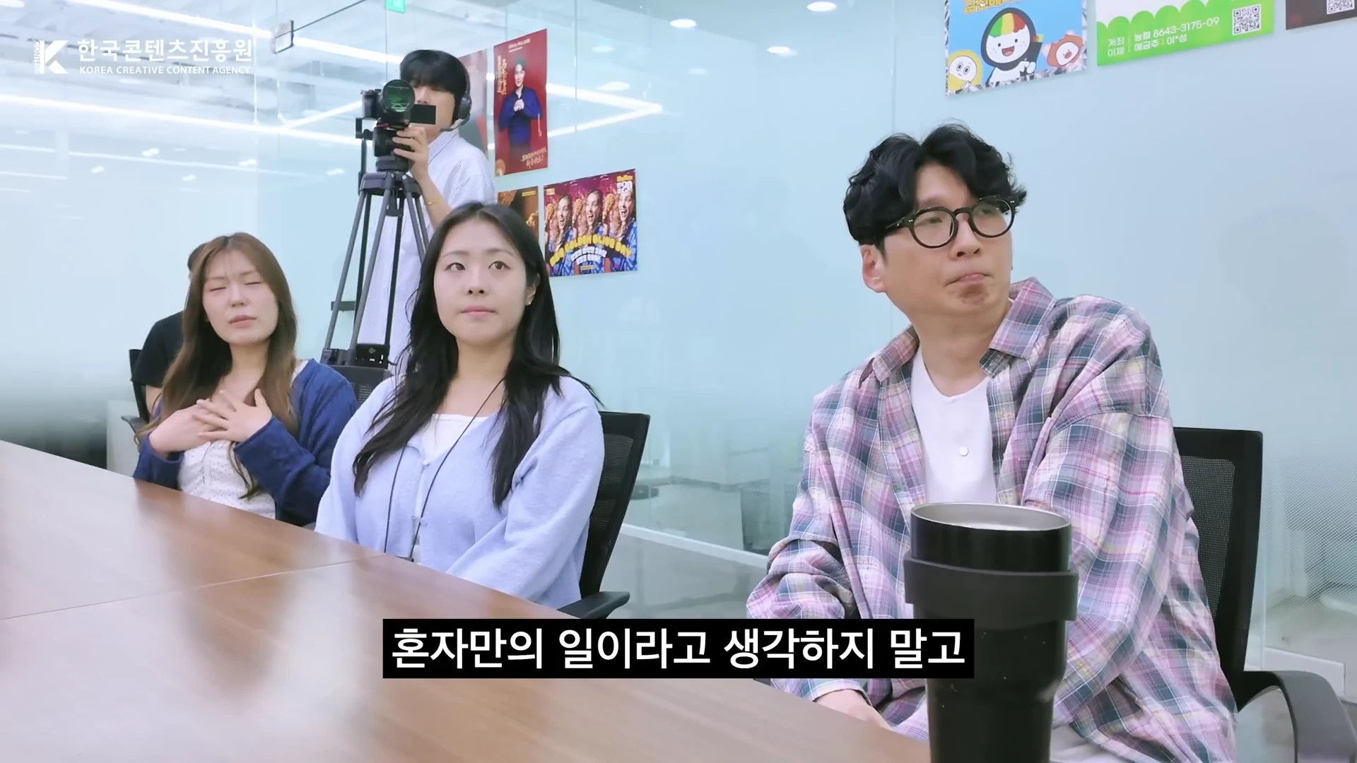 사무실에서 세 명의 출연진이 앉아 이야기를 나누고 뒤편에는 촬영 장비와 포스터가 보인다. 화면 하단에는 자막이 있다.