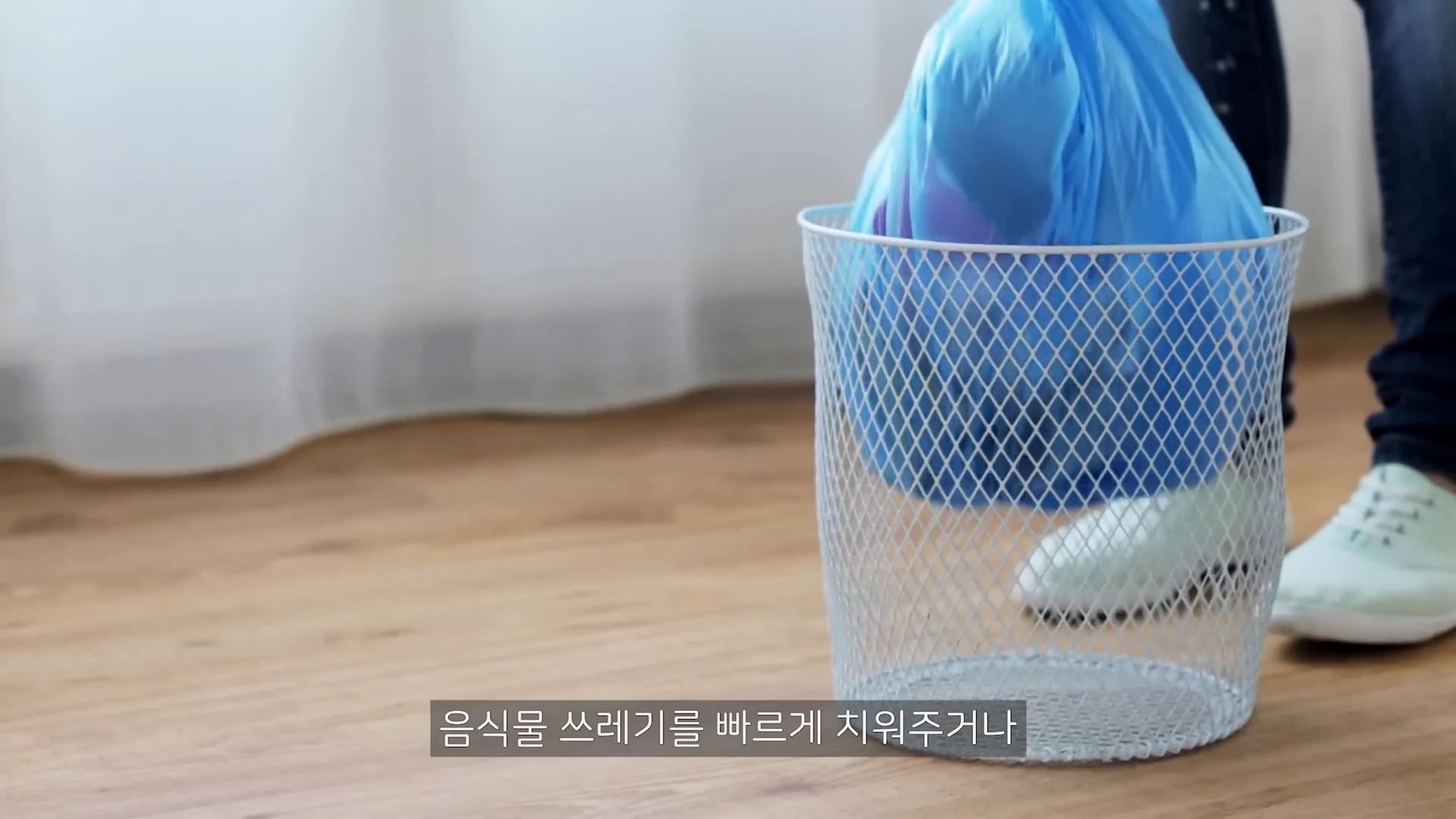 흰색 금속 망사 쓰레기통에 파란색 비닐봉지가 들어있고 한 사람이 옆에 서 있는 장면