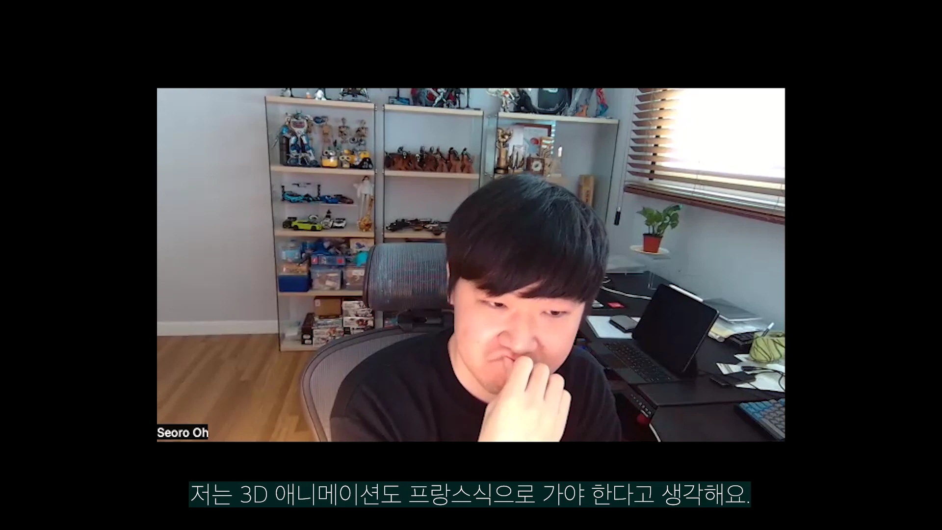 한 남성이 자신의 작업실로 보이는 공간에서 의자에 앉아 고민하는 듯한 표정으로 손을 입가에 대고 있으며, 배경에는 다양한 피규어와 장식품이 진열된 선반이 보입니다. 화면 하단에는 '저는 3D 애니메이션도 프랑스식으로 가야 한다고 생각해요.'라는 자막이 표시되어 있습니다.