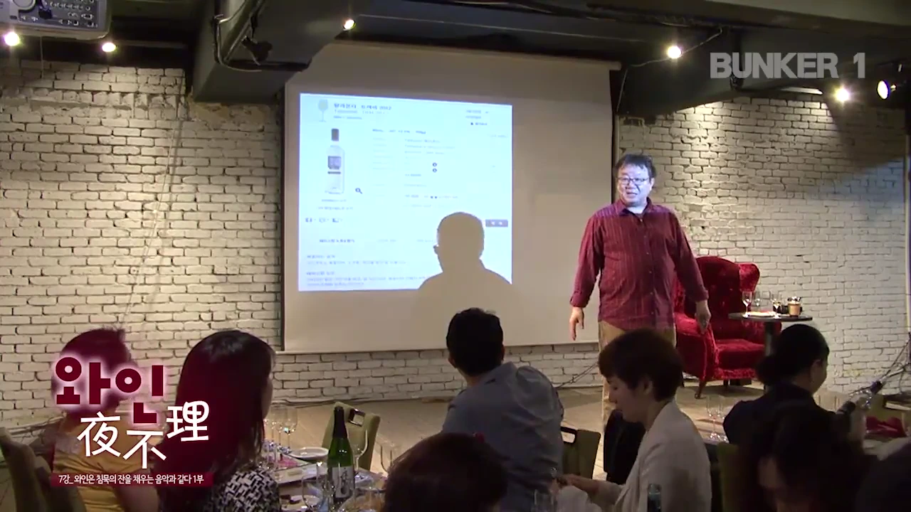 실내 강연장에서 안경을 쓴 중년 남성이 스크린에 띄워진 와인 정보를 가리키며 청중에게 설명하고 있고, 앞에는 와인잔이 놓인 테이블들이 배치되어 있다.