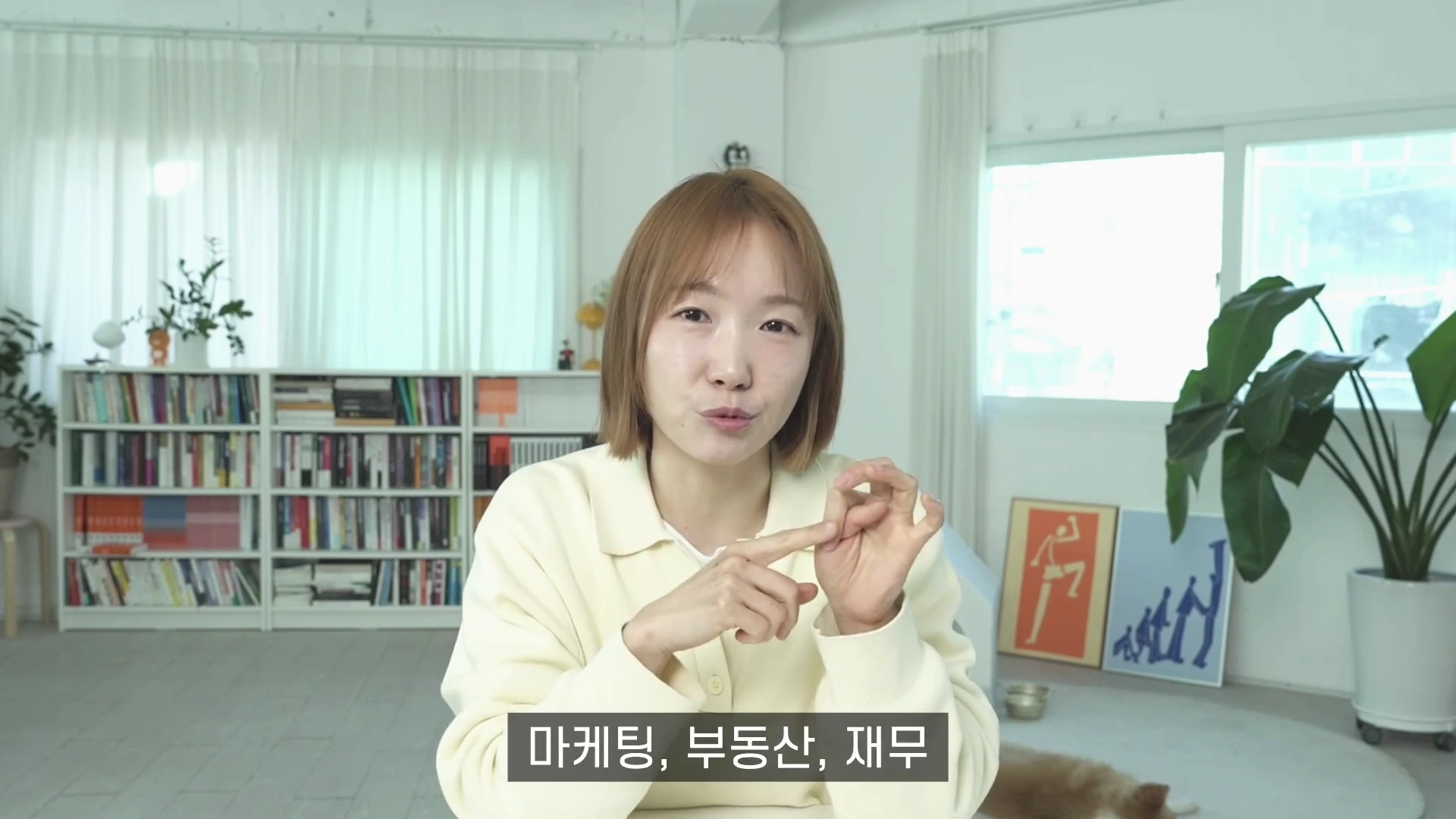 단발머리를 한 여성이 밝은색 가디건을 입고 밝은 실내에서 손가락을 사용해 숫자를 세며 이야기를 하고 있으며, 하단에는 '마케팅, 부동산, 재무'라는 자막이 표시되어 있습니다.