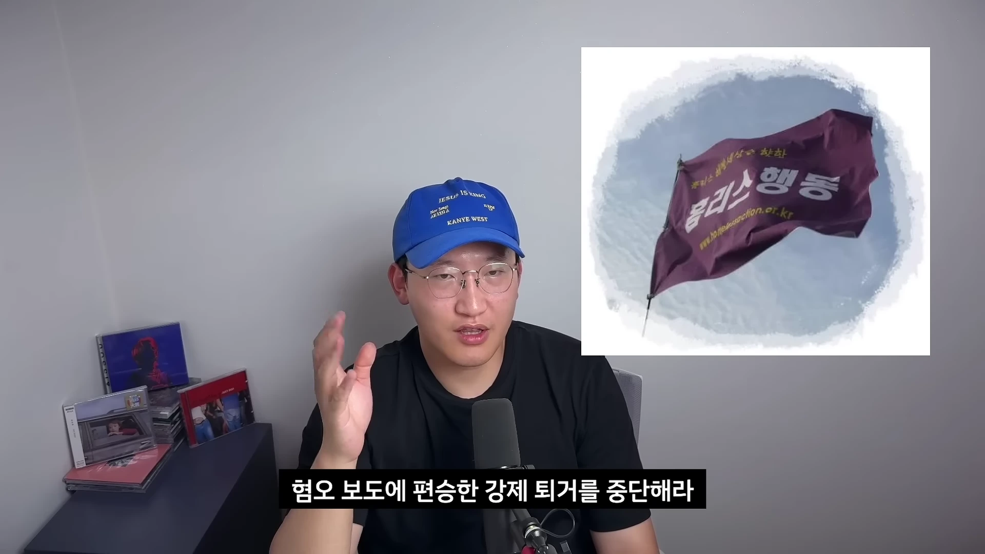파란색 모자를 쓴 남성이 마이크 앞에 앉아 말하고 있고, 화면 오른쪽 상단에는 '홈리스행동'이라는 글자가 적힌 보라색 깃발 이미지가 합성되어 있습니다.