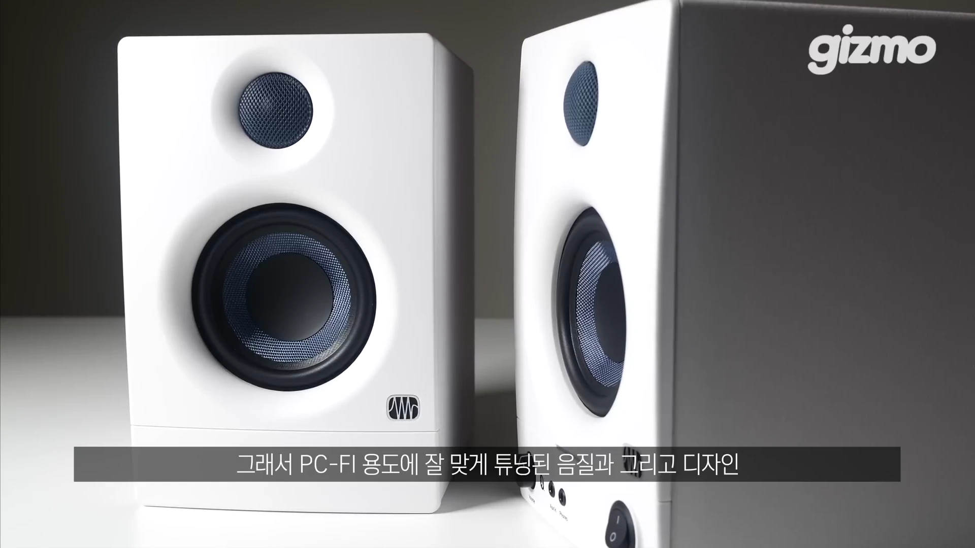흰색 모니터 스피커 한 쌍이 책상 위에 놓여 있으며, 하단에 로고와 조작 버튼이 보이고 화면 하단에는 자막이 있습니다.