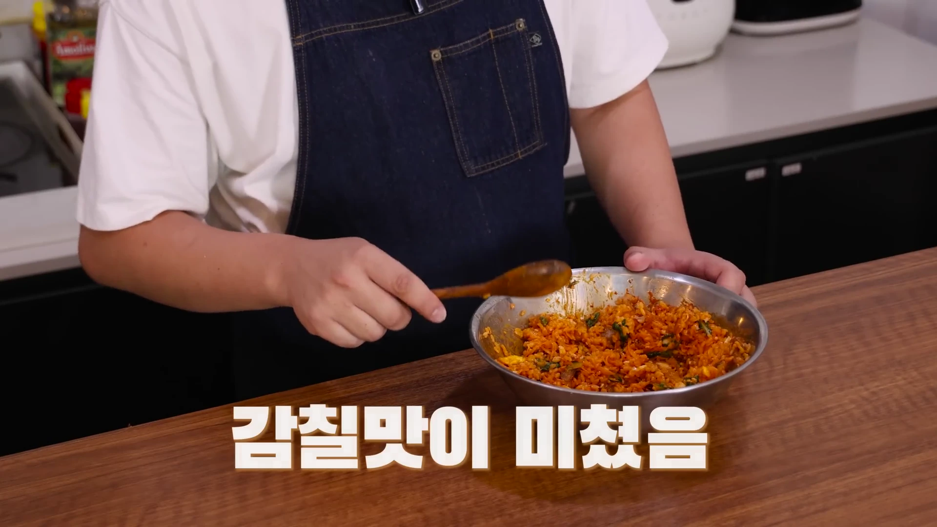 앞치마를 두른 요리사가 나무 숟가락으로 양푼에 담긴 붉은색 비빔밥을 비비고 있는 모습이 담긴 영상 캡처 화면이며, 화면 하단에는 흰색 글씨로 '감칠맛이 미쳤음'이라는 자막이 적혀 있다.