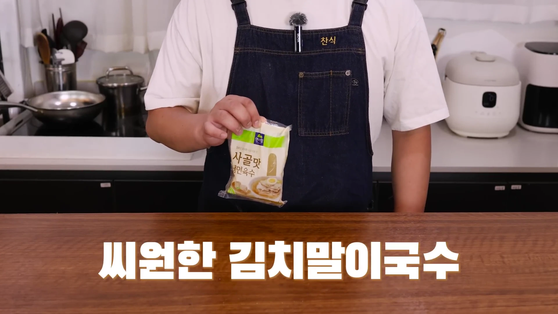 앞치마를 두른 요리사가 한 손으로 냉면 육수 팩을 들고 있고 화면 하단에는 '씨원한 김치말이국수'라는 자막이 크게 적혀 있습니다.
