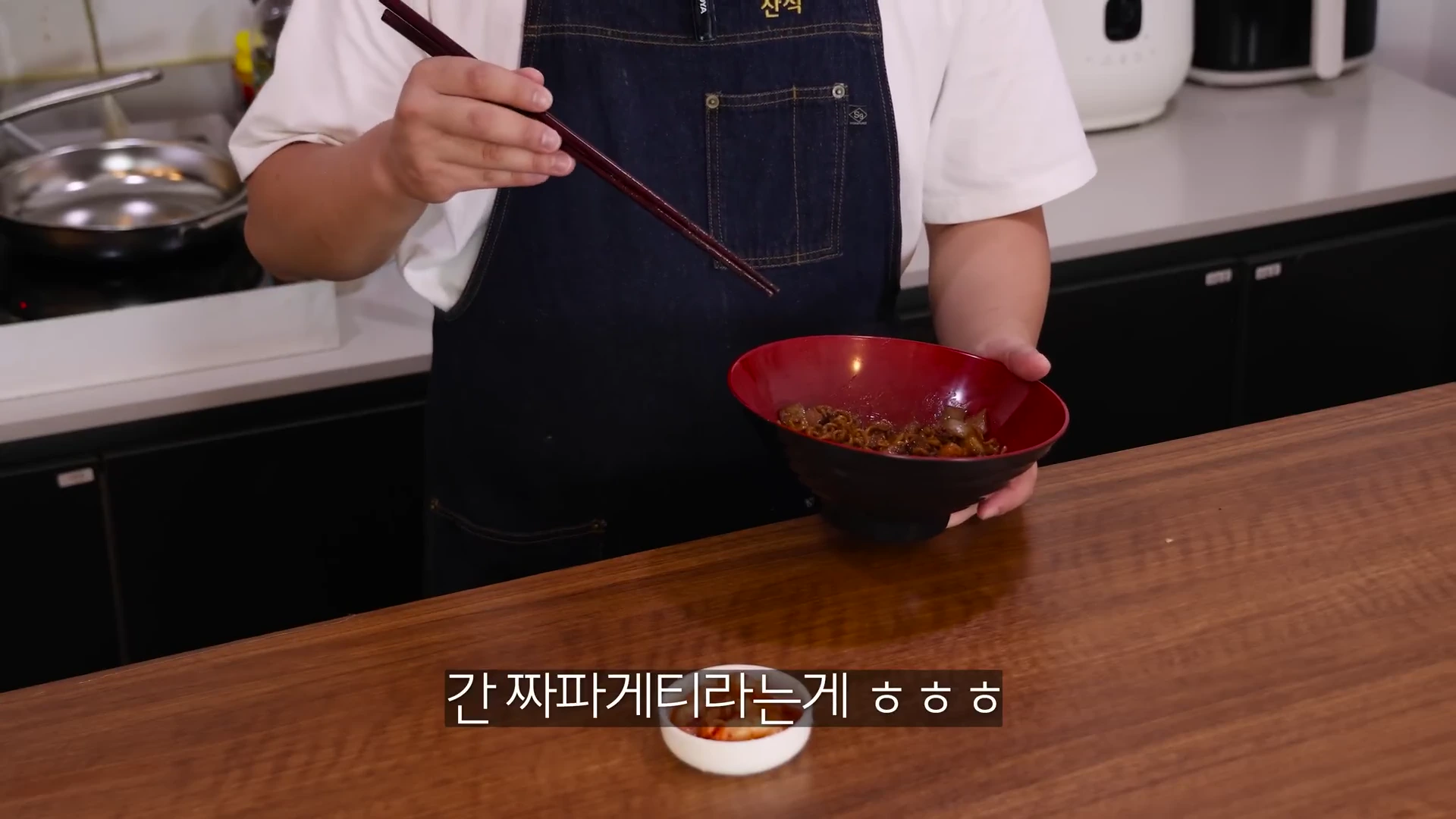 앞치마를 두른 요리사가 붉은색 면기에 담긴 짜장면 요리를 들고 젓가락을 든 채 서 있는 모습