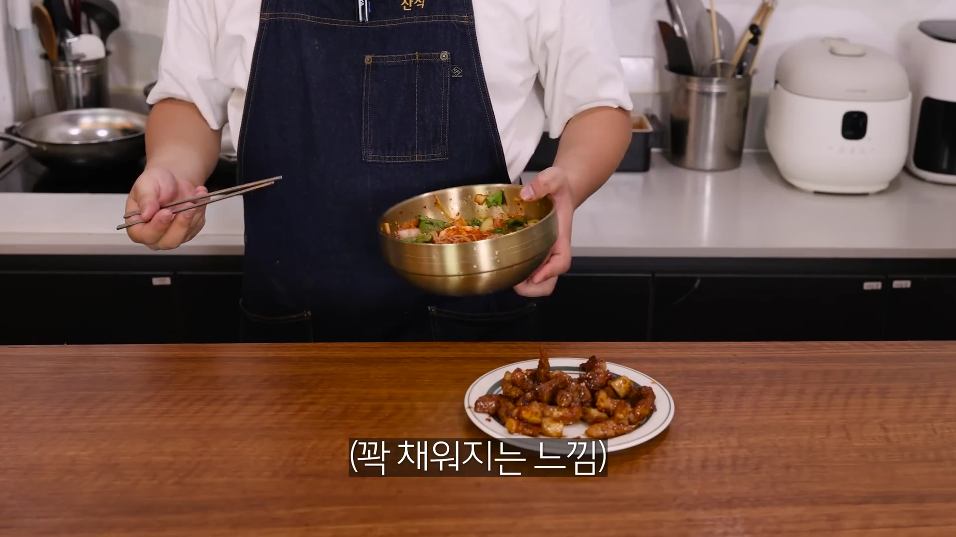 앞치마를 두른 요리사가 한 손에는 놋그릇에 담긴 비빔국수를 들고 다른 한 손에는 젓가락을 든 채 조리대 앞에 서 있고 앞쪽 접시에는 양념된 고기가 놓여 있다.