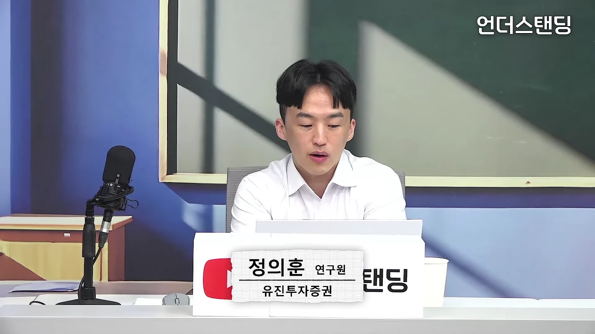 정장을 입은 남성 출연자가 책상 앞에 앉아 이야기하고 있고, 앞쪽에는 이름과 소속이 적힌 명패와 검은색 스탠드 마이크가 놓여 있습니다.