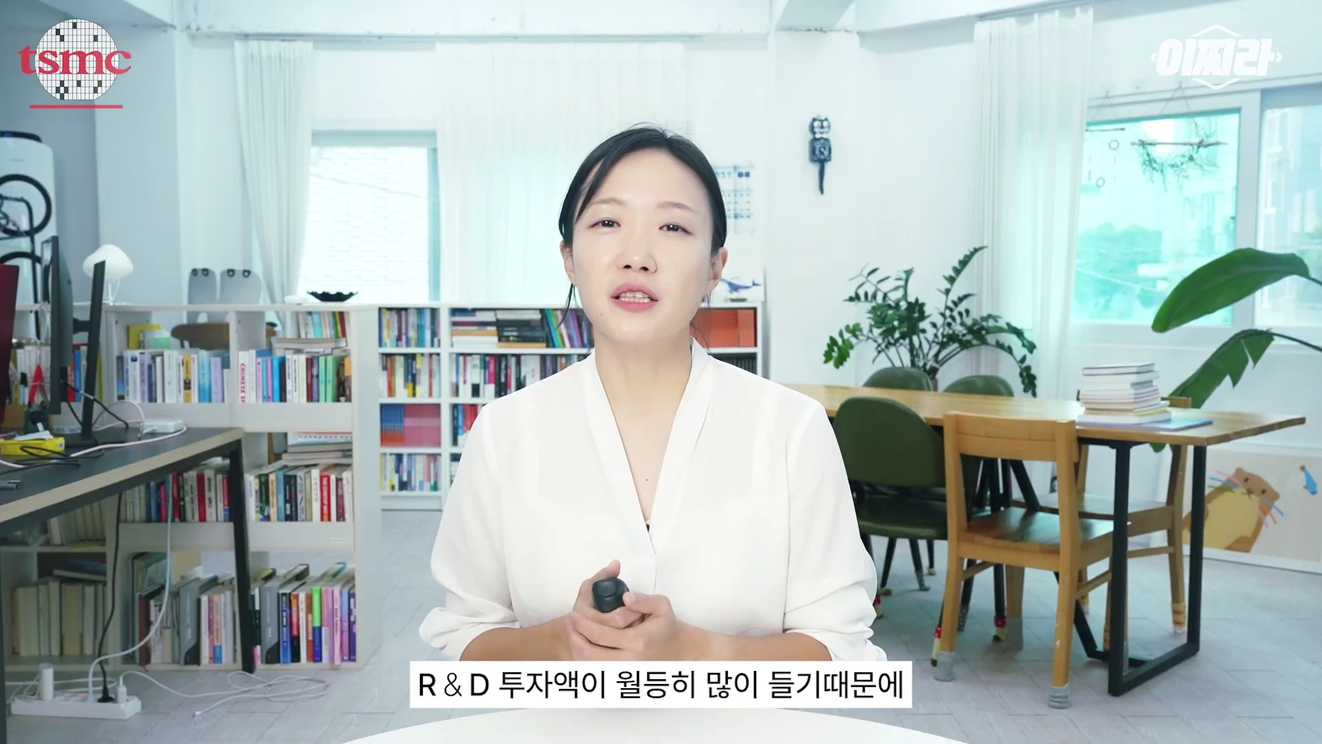 흰색 블라우스 차림의 여성이 밝은 서재를 배경으로 서서 카메라를 바라보고 있으며, 화면 하단에는 'R & D 투자액이 월등히 많이 들기 때문에'라는 자막이 표시되어 있다.
