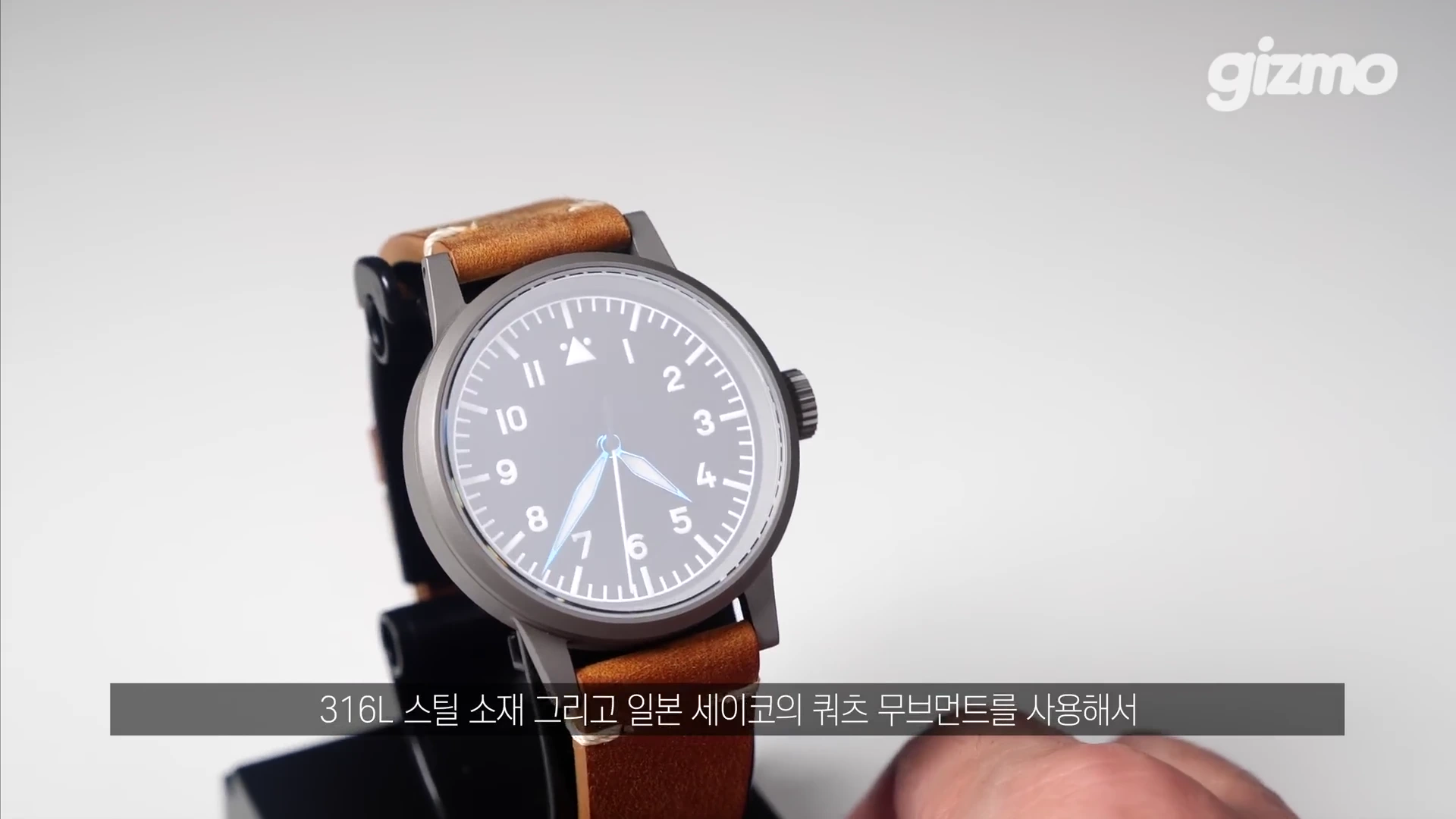 흰색 배경 위에 갈색 가죽 스트랩이 달린 검은색 다이얼의 군용 스타일 손목시계가 스탠드에 거치되어 있다. 화면 하단에는 제품 사양을 설명하는 한국어 자막이 표시되어 있다.