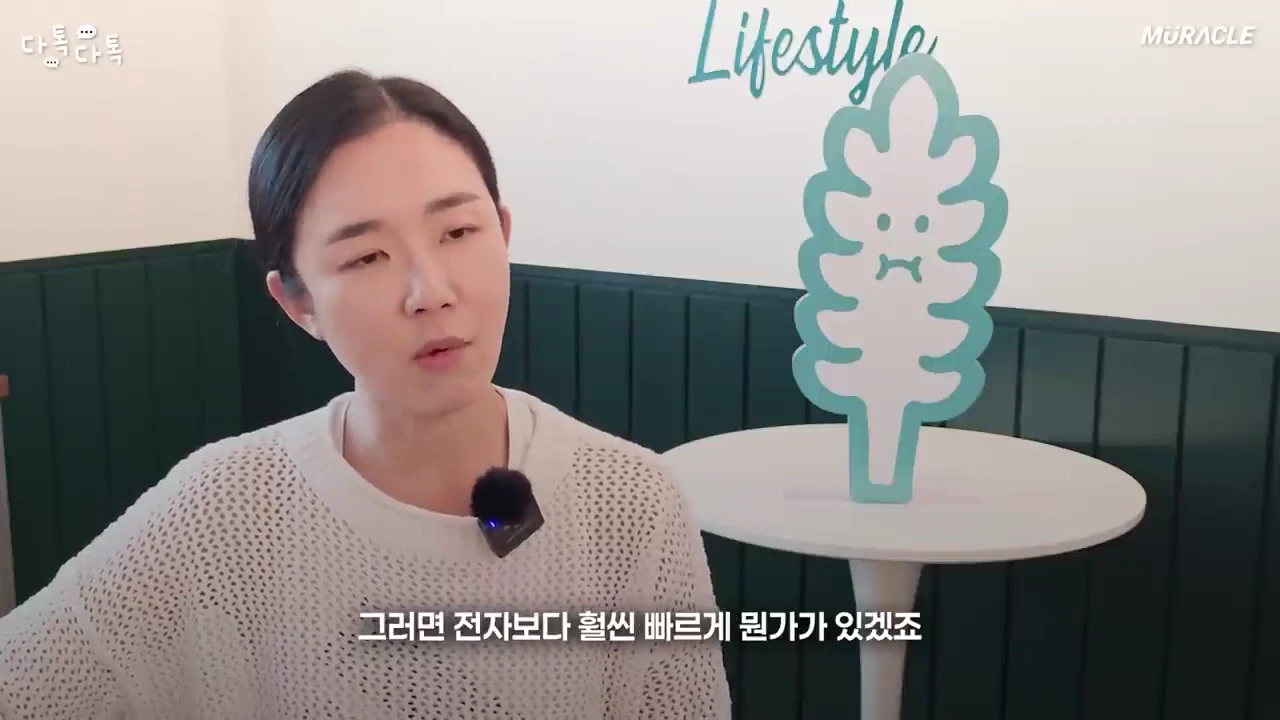 흰색 니트를 입은 여성이 밝은 실내에서 이야기를 하고 있으며 옆에는 잎사귀 모양의 캐릭터 조형물이 놓여 있습니다.