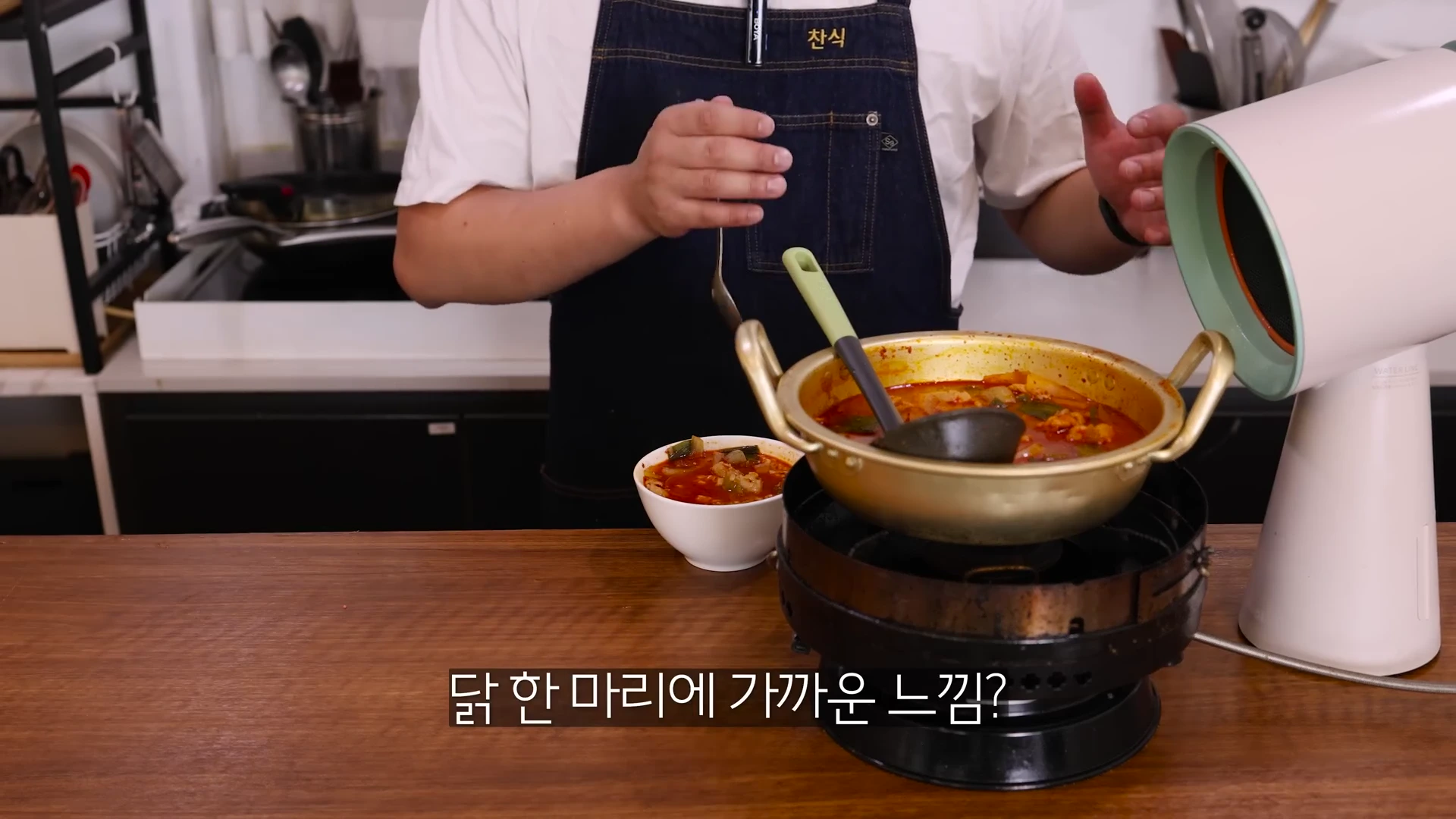 앞치마를 두른 요리사가 주방에서 냄비에 담긴 얼큰한 국물 요리를 설명하고 있고 옆에는 이동식 미니 후드가 놓여 있다.