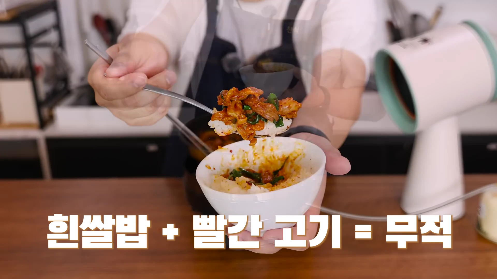 요리사가 숟가락으로 매콤한 양념에 볶은 고기와 부추를 흰쌀밥 위에 얹어 들어 보이고 있으며, 화면 하단에는 흰색 자막이 적혀 있습니다.