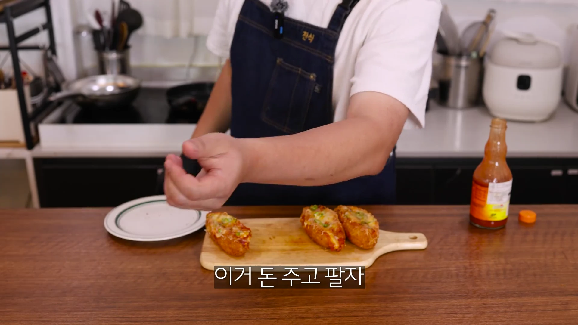 앞치마를 두른 요리사가 도마 위에 올려진 청양고추가 토핑된 핫도그를 가리키고 있으며, 옆에는 핫소스 병이 놓여 있다.