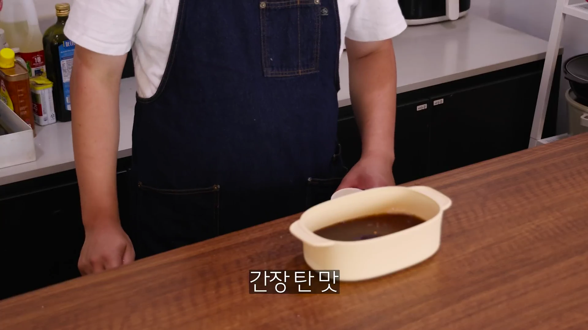 앞치마를 두른 요리사가 주방 조리대 위에 놓인 타원형 용기에 배 음료와 간장 등을 섞어 둔 모습을 상반신 위주로 담은 장면.