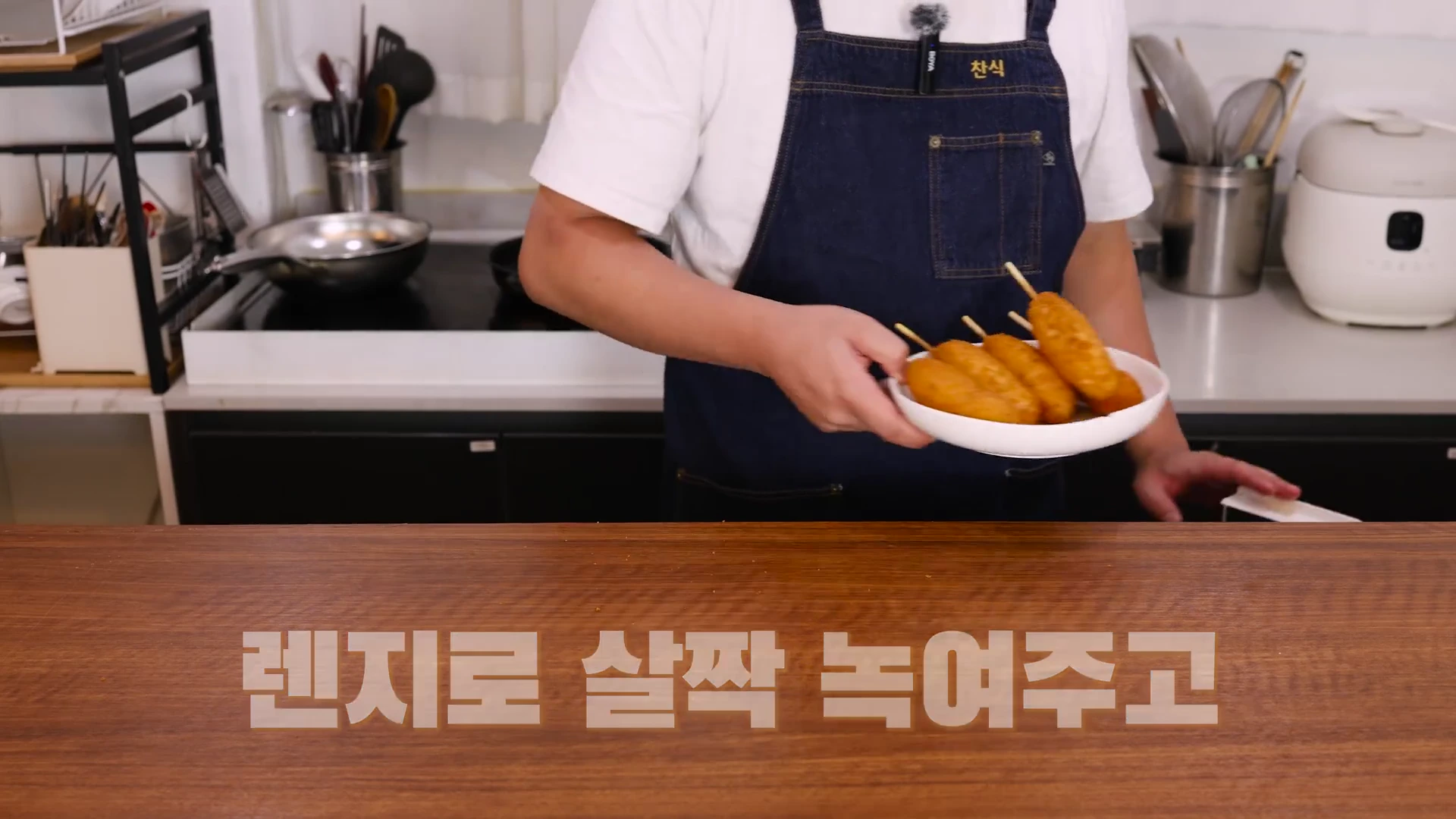 앞치마를 두른 남성이 전자레인지에 해동한 핫도그가 담긴 접시를 손에 들고 주방 조리대 앞에 서 있는 모습.