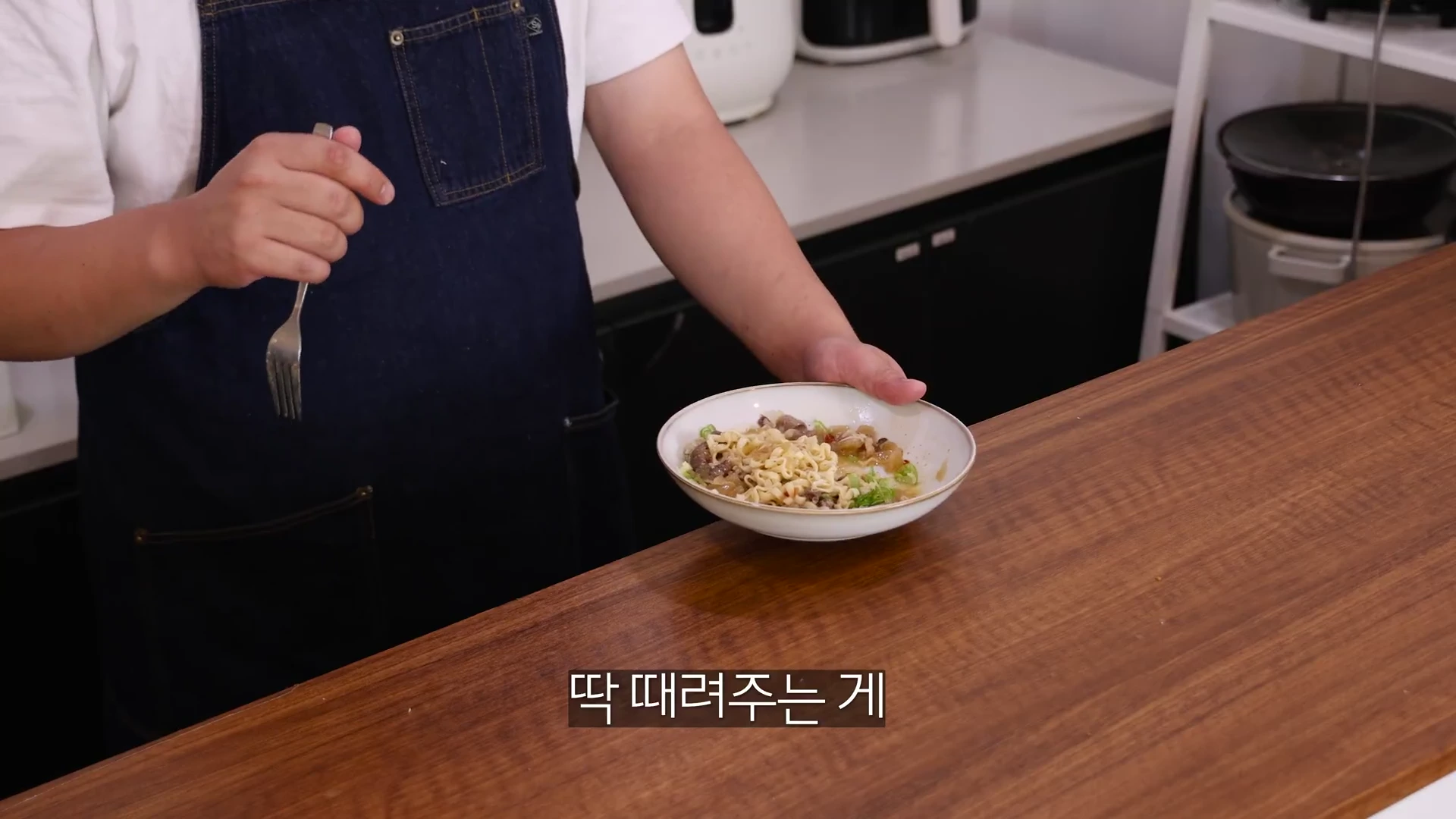 주방에서 앞치마를 두른 요리사가 포크를 들고 완성된 면 요리가 담긴 그릇을 보여주고 있다.
