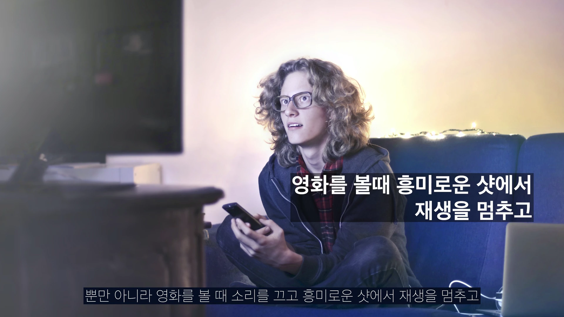 안경을 쓴 곱슬머리 청년이 어두운 방 안 소파에 앉아 TV 화면을 집중해서 바라보며 리모컨을 들고 있는 모습.