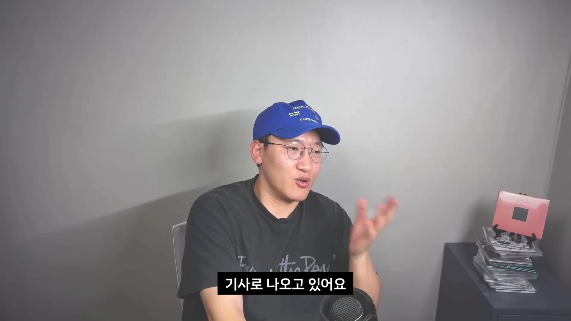 파란색 모자를 쓰고 안경을 쓴 남성이 마이크 앞에서 손을 움직이며 이야기하고 있다.