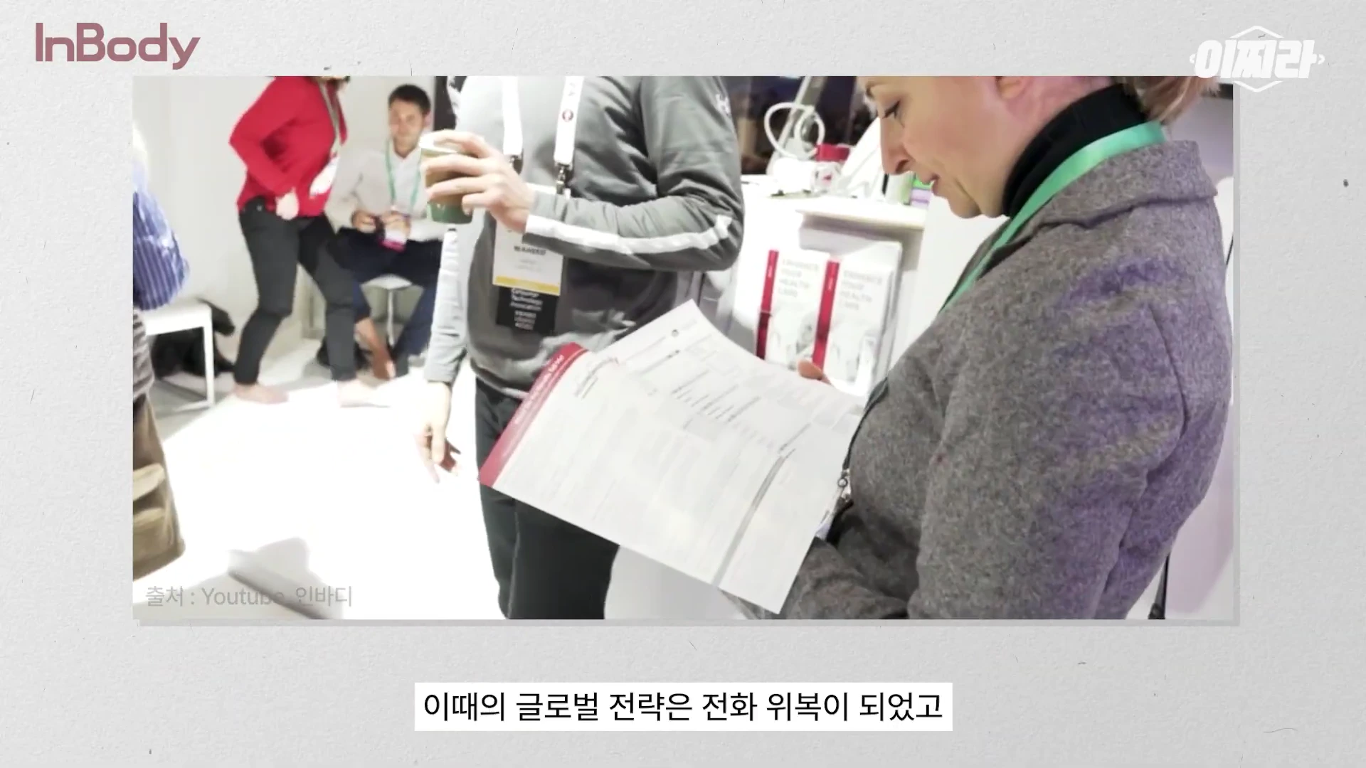 실내 전시장에서 서류를 살펴보고 있는 여성과 그 옆에서 음료를 들고 대화하는 참가자들의 모습이 담긴 영상 장면.