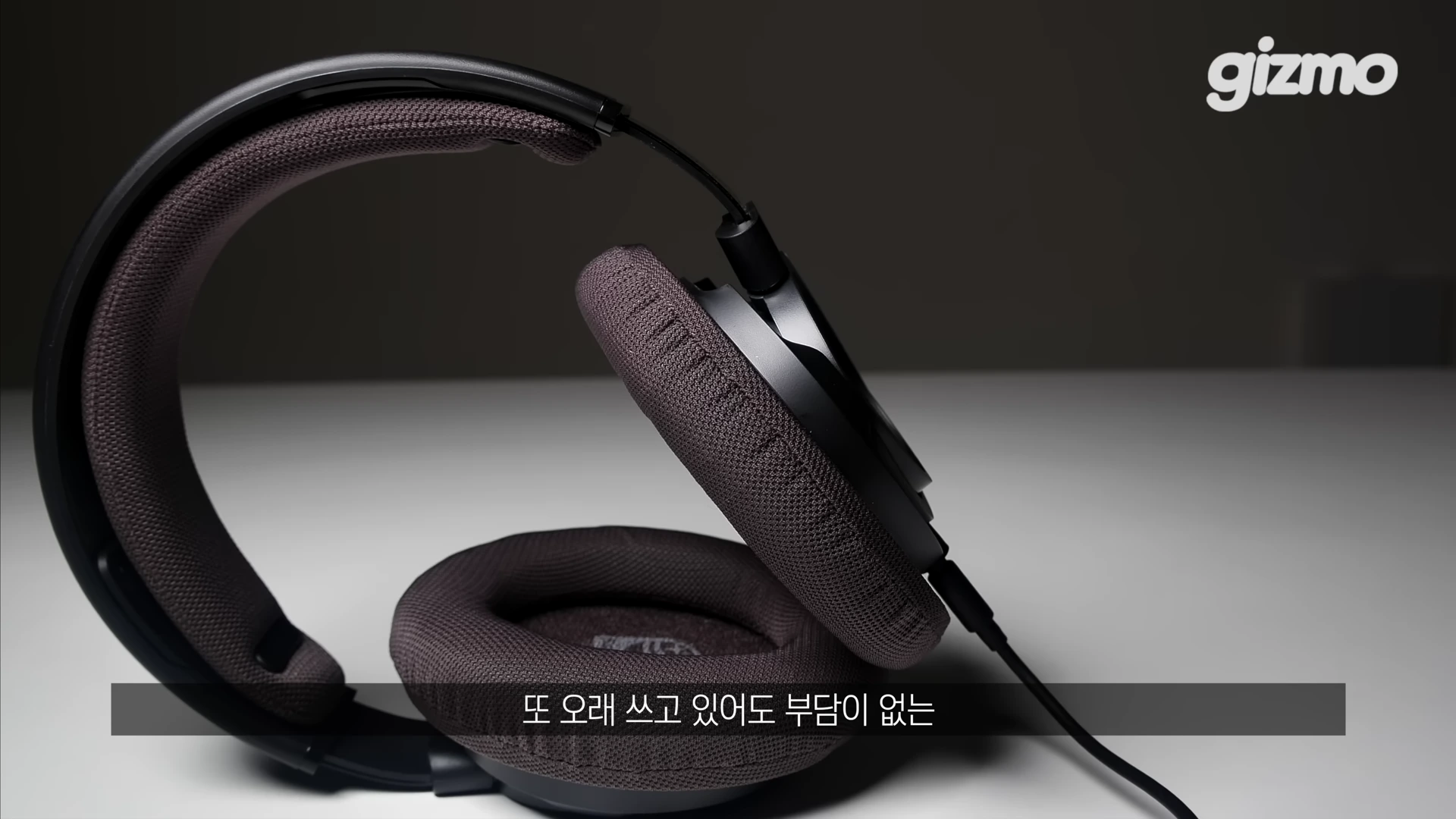 짙은 회색 패브릭 소재의 이어패드와 헤드밴드가 장착된 유선 헤드폰이 하얀 테이블 위에 놓여 있는 모습.