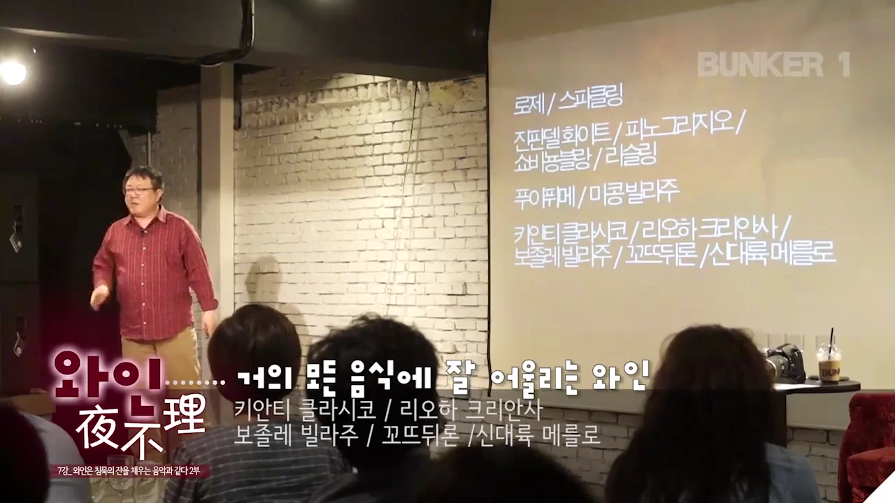 강연자가 화면 앞에 서서 와인 종류가 나열된 슬라이드를 가리키며 청중에게 설명하고 있는 모습